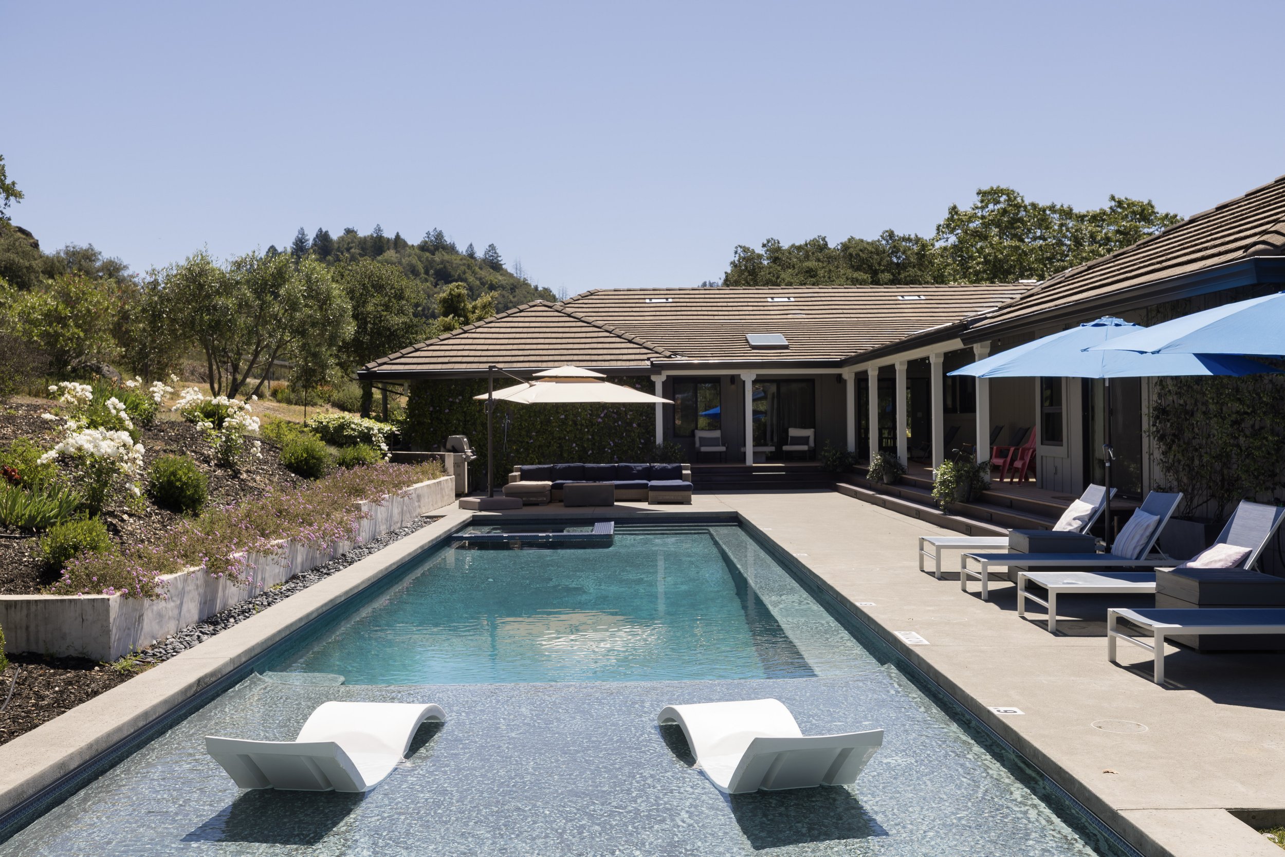 Backyard with a rectangular swimming pool, lounge chairs, umbrellas, and a house with a tiled roof, surrounded by trees and hills under a clear blue sky.
