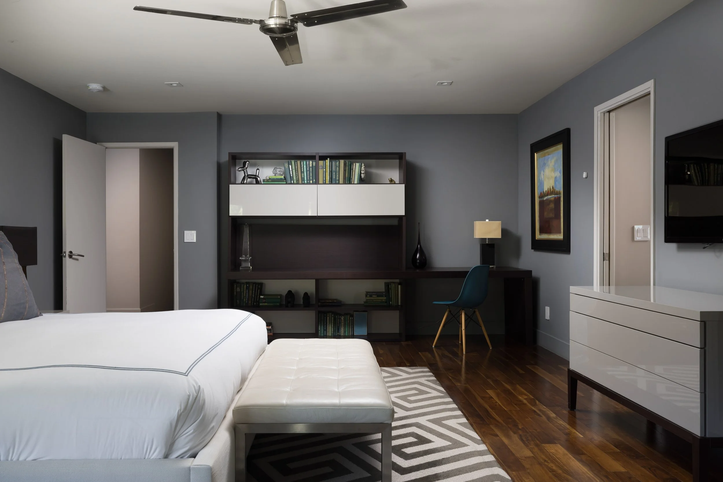 Interior of a modern bedroom with gray walls, a large bed with white linens, a beige ottoman, and a black and white bookshelf with decorative items. The room has a ceiling fan, a painting on the wall, and a dresser with a flat-screen TV.