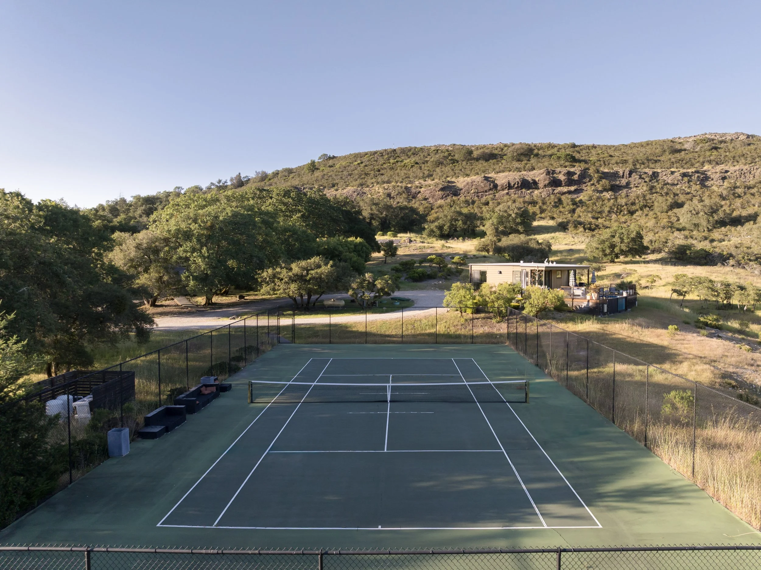 A tennis court surrounded by a black chain-link fence with residential buildings in the background, hills covered with trees and rocky outcroppings, and a clear blue sky overhead.