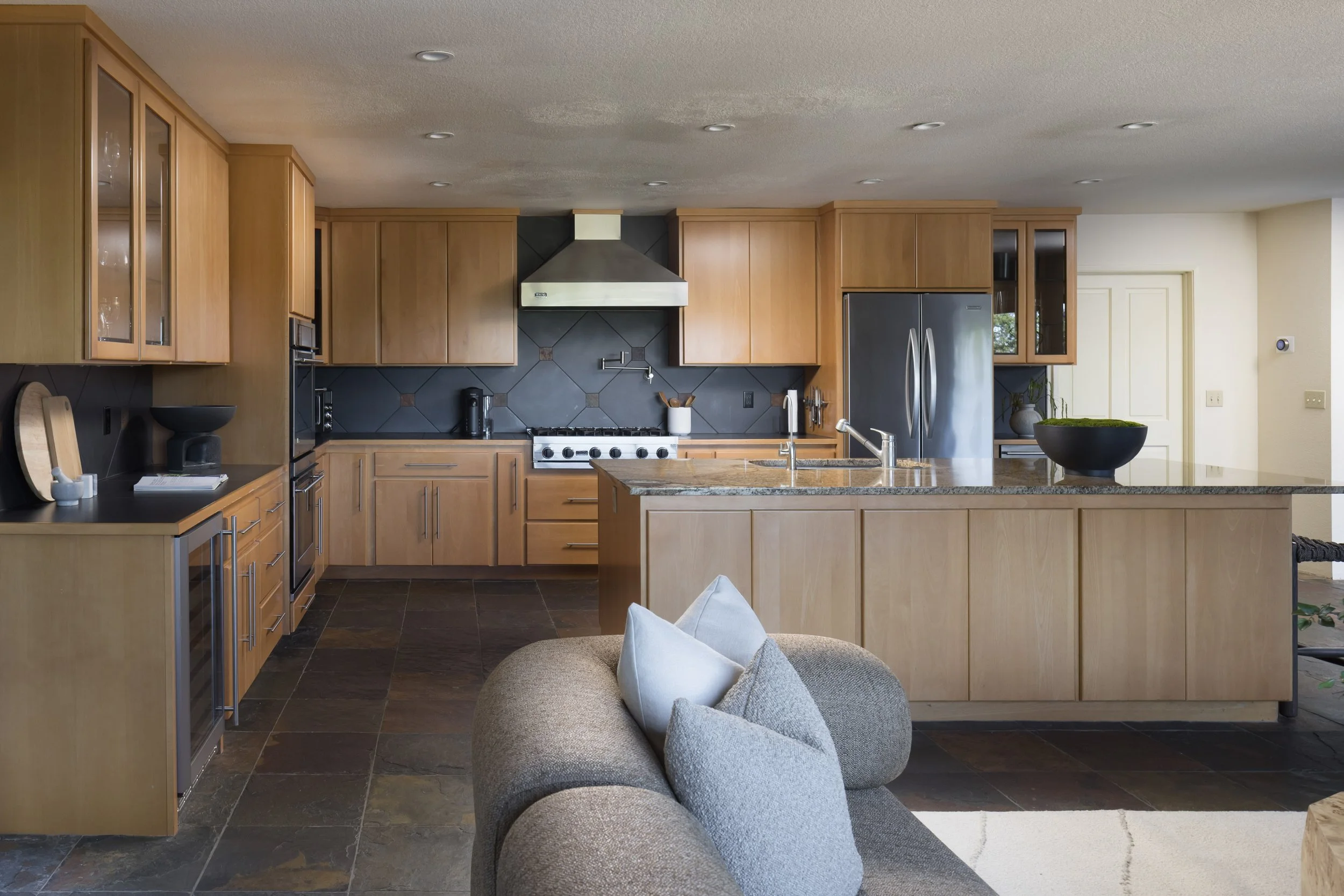 Modern kitchen with wooden cabinets, stainless steel appliances, black tile backsplash, and a large kitchen island with a sink and decorative bowl.