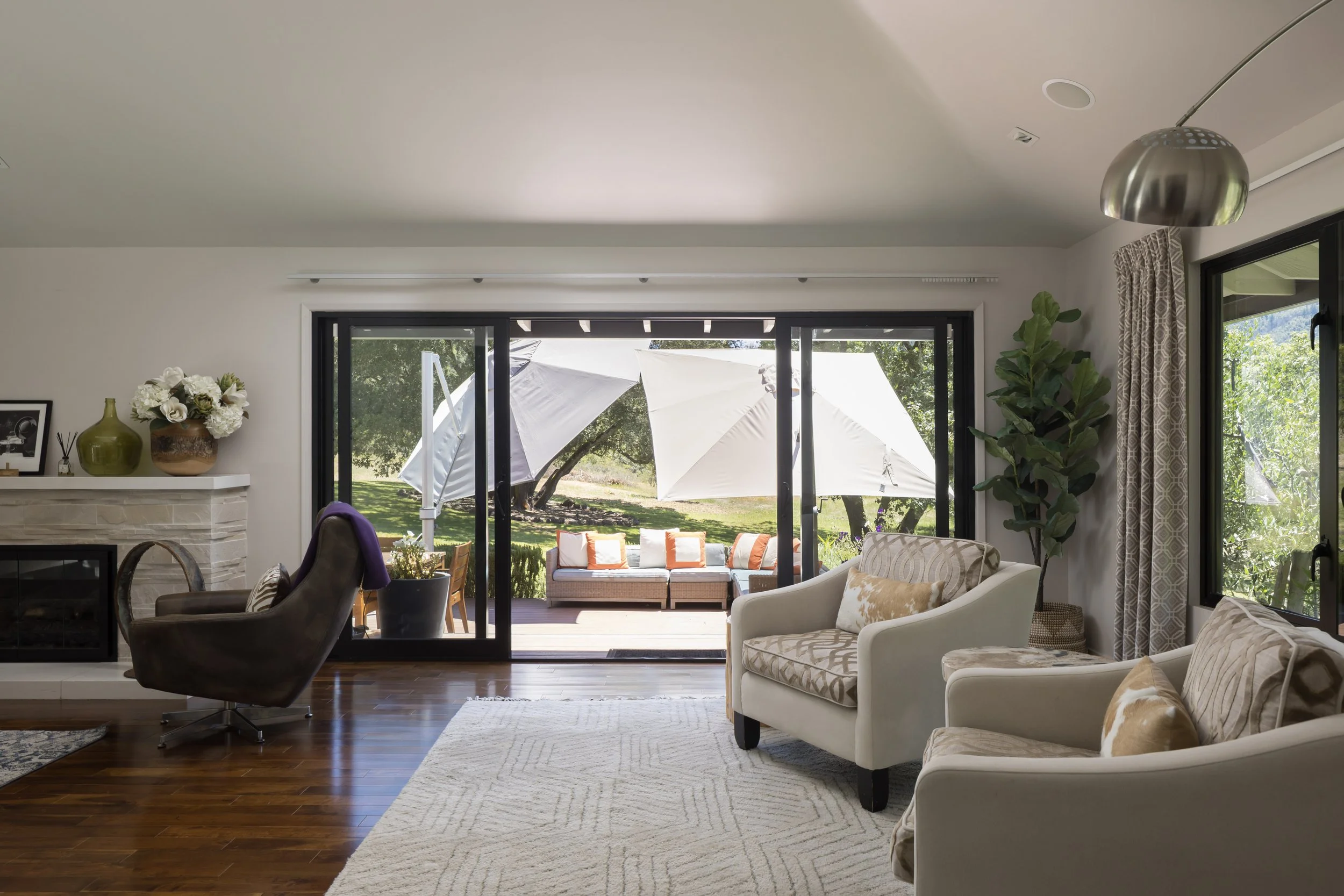 Living room with white couches and a large sliding glass door opening to an outdoor patio with umbrellas and trees.
