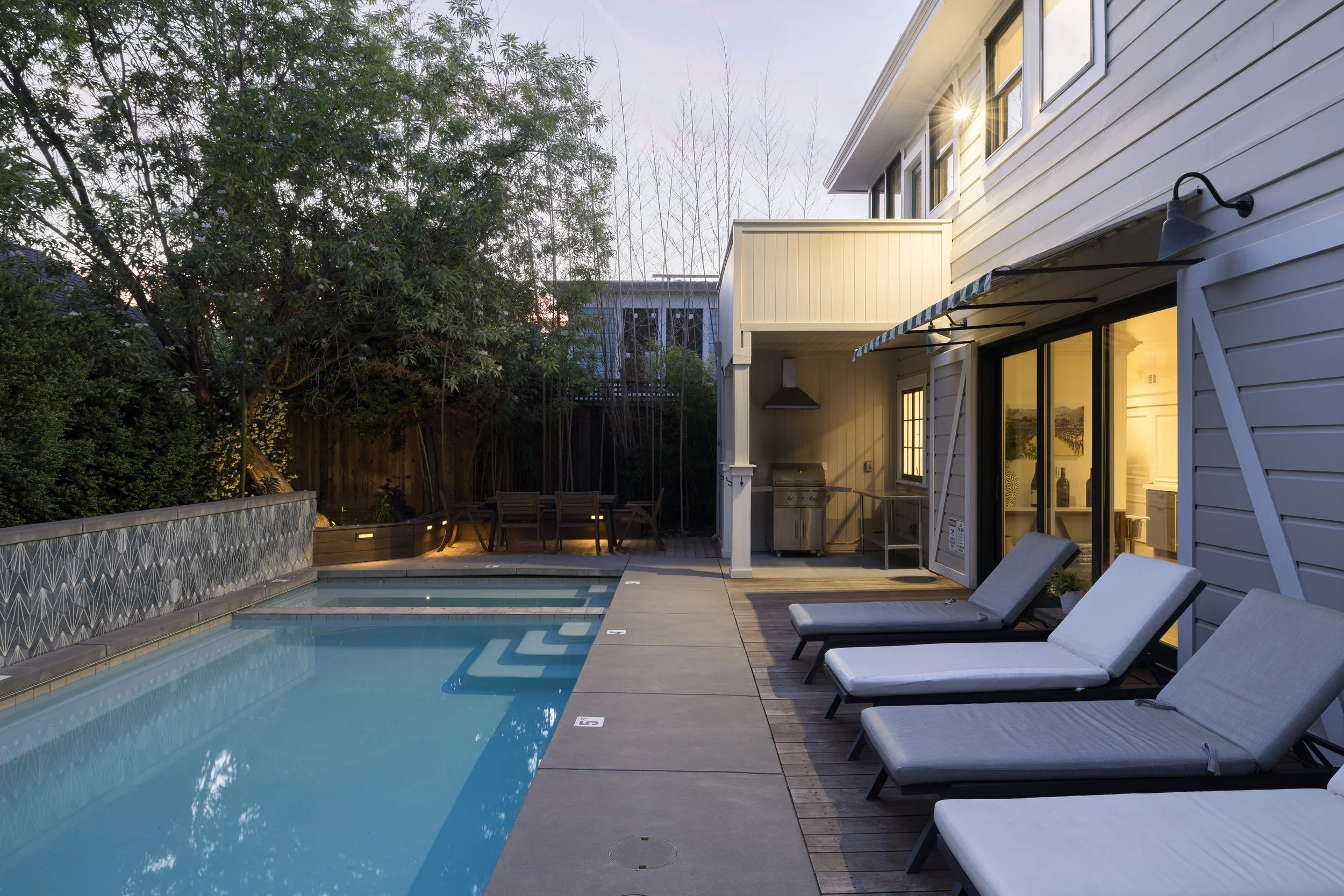 Backyard view with a swimming pool, lounge chairs, outdoor dining table, and a house with a sliding glass door and outdoor kitchen area, with trees and a fence in the background.
