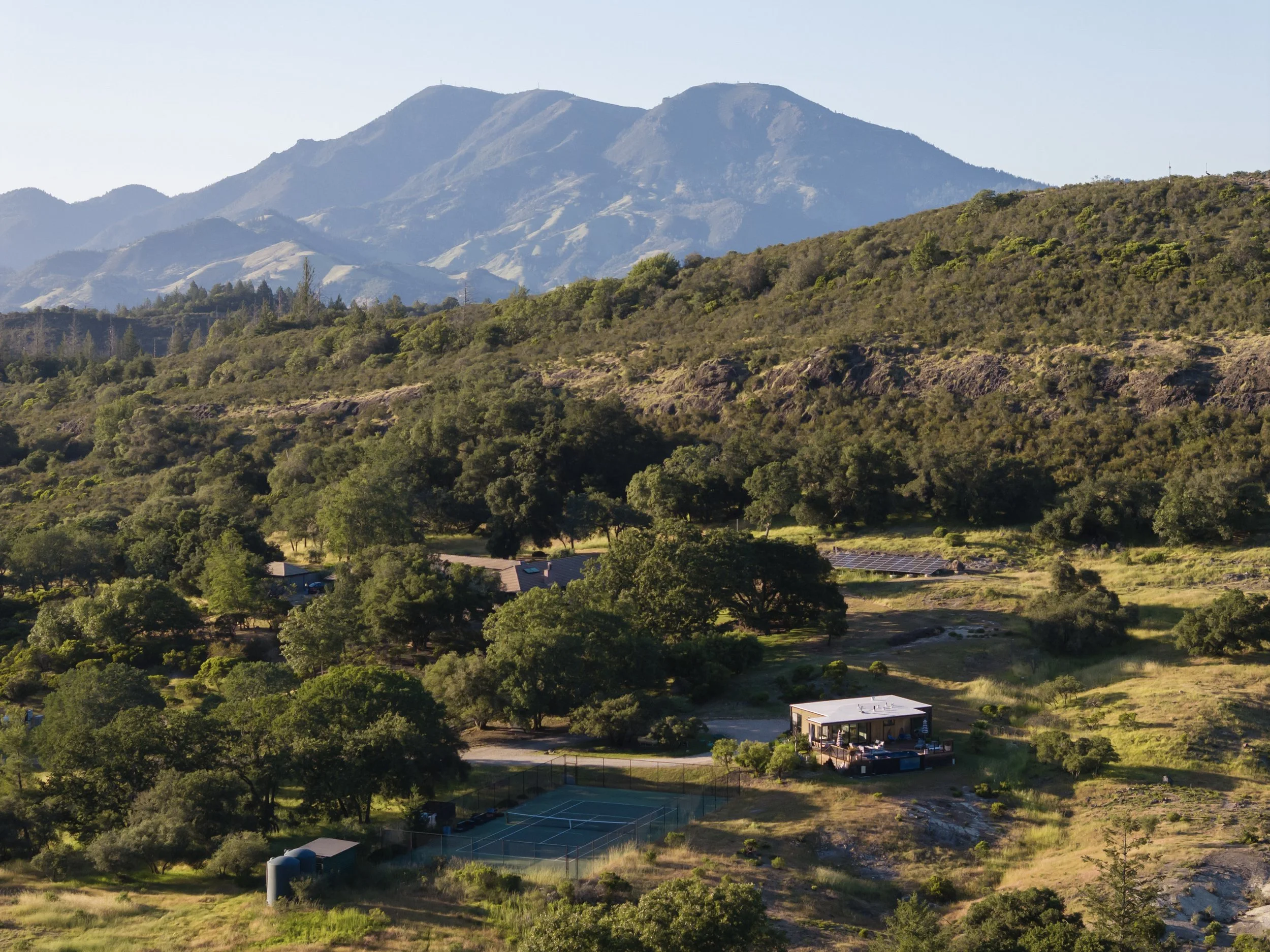 A view of green hills and mountains in the distance with a residential house, tennis courts, and trees in the foreground.