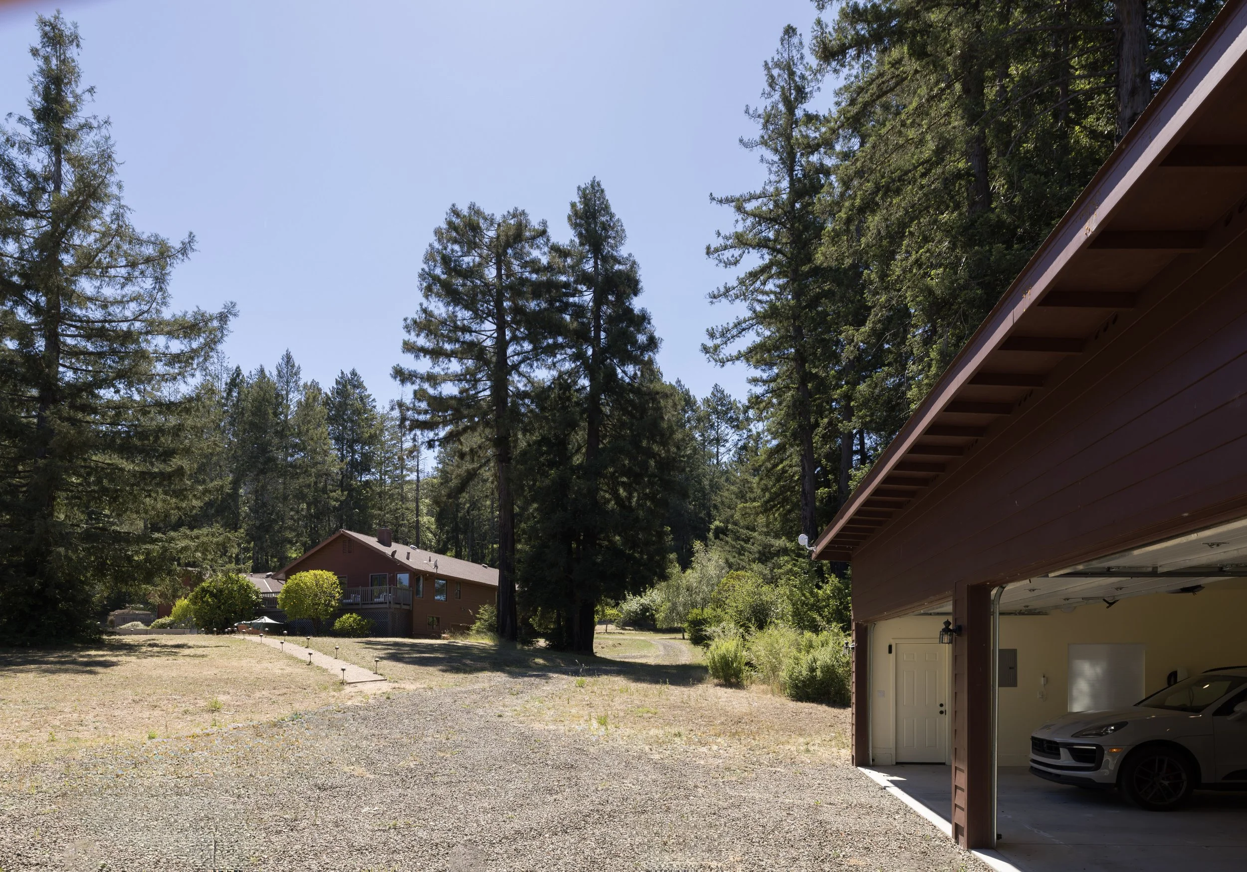 View of a backyard with tall trees, a neighboring house, and a portion of a garage with a car inside, under a clear blue sky.
