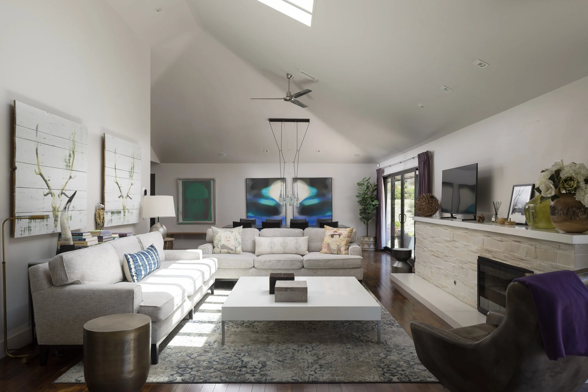 A spacious living room with a large white sectional sofa adorned with various decorative pillows, a dark brown leather armchair with a purple blanket, a white coffee table with a abstract sculpture, a beige rug with blue and white patterns, a white b