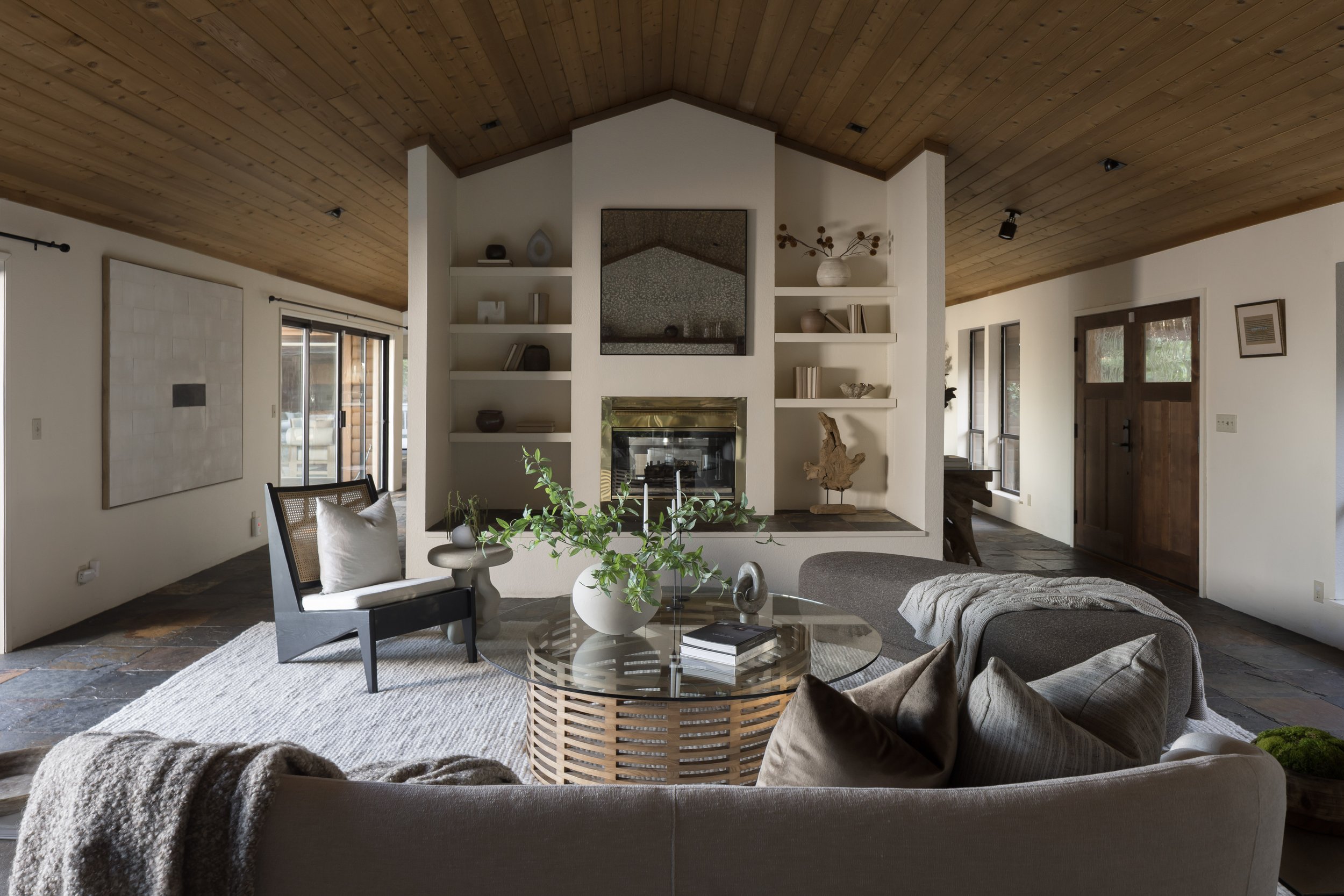 Living room with wooden ceiling, fireplace, built-in shelves, sliding glass door, and modern furniture including a sofa, armchair, and coffee table with plants.