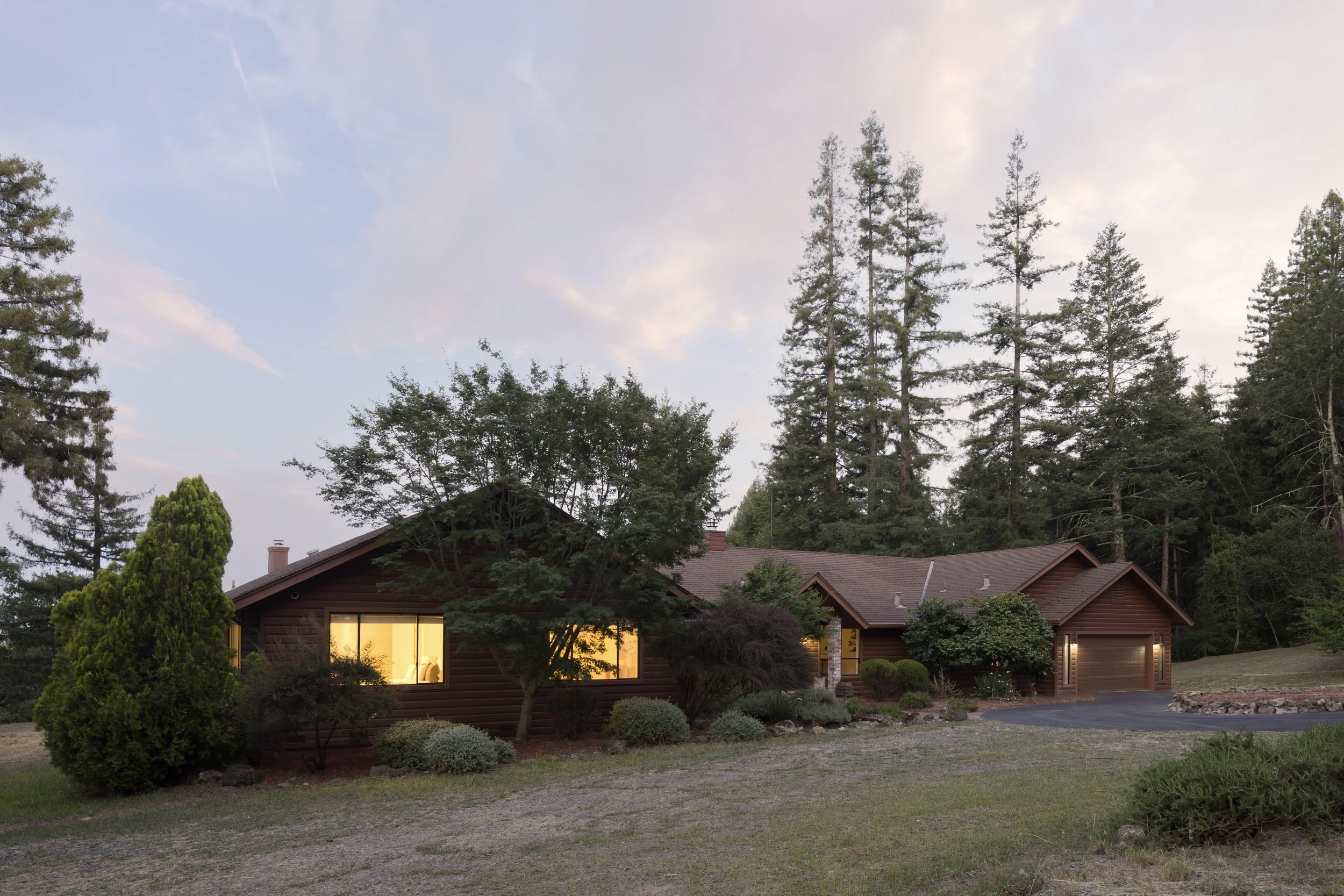 A suburban house with a brown exterior and large windows at sunset. The house is surrounded by tall trees and trimmed shrubs. The sky is partly cloudy, with a soft pink hue from the setting sun.
