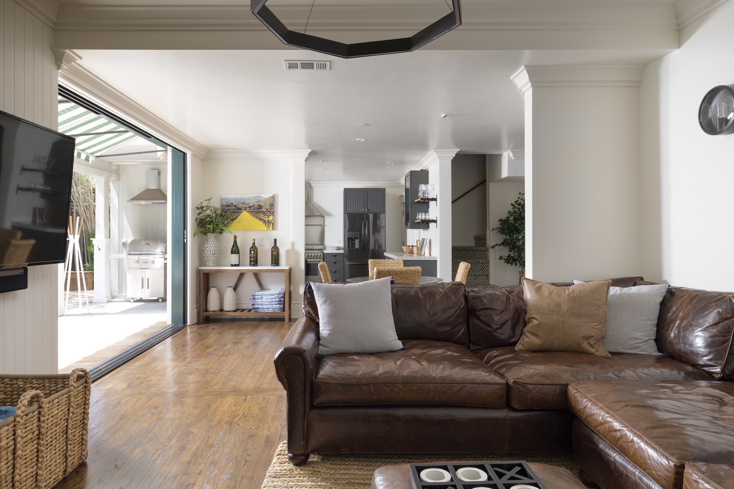 Living room with brown leather sofa, gray and white pillows, wooden coffee table, and sliding glass door leading outside. Kitchen area visible in the background with black refrigerator, stove, and a painting on the wall. Indoor plants and decorative 