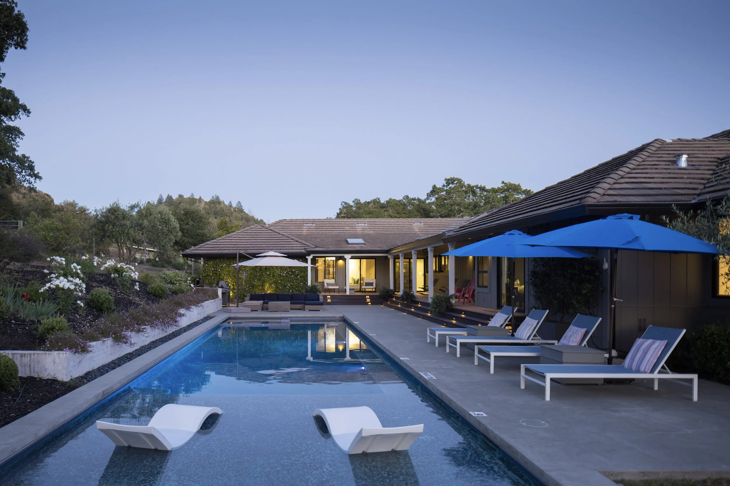Backyard with swimming pool, outdoor seating, umbrellas, and a house with large windows at dusk.
