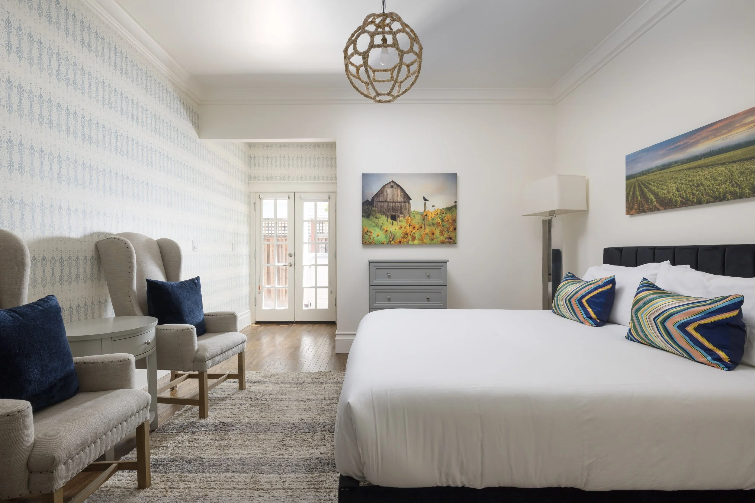 A bedroom with a white bed, colorful striped pillows, and artwork of farmland and a barn on the wall. There are beige armchairs with dark blue pillows and a small round table in front. A dresser and a floor lamp are also visible, with a door leading 