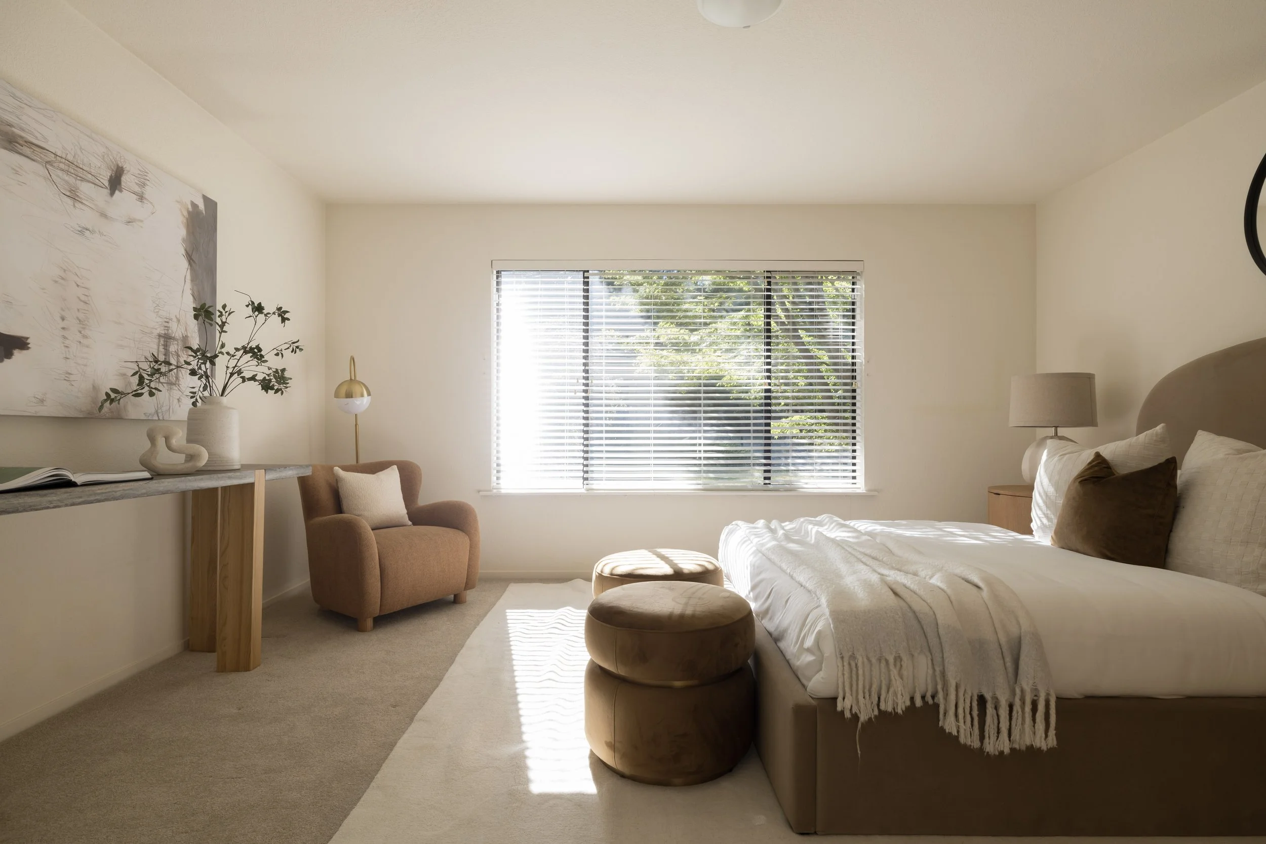 A bedroom with a large window with blinds, a bed with beige bedding and pillows, a bedside table with a lamp, a brown armchair with a pillow, an abstract artwork on the wall, a console table with decorative objects, and two matching round ottomans at