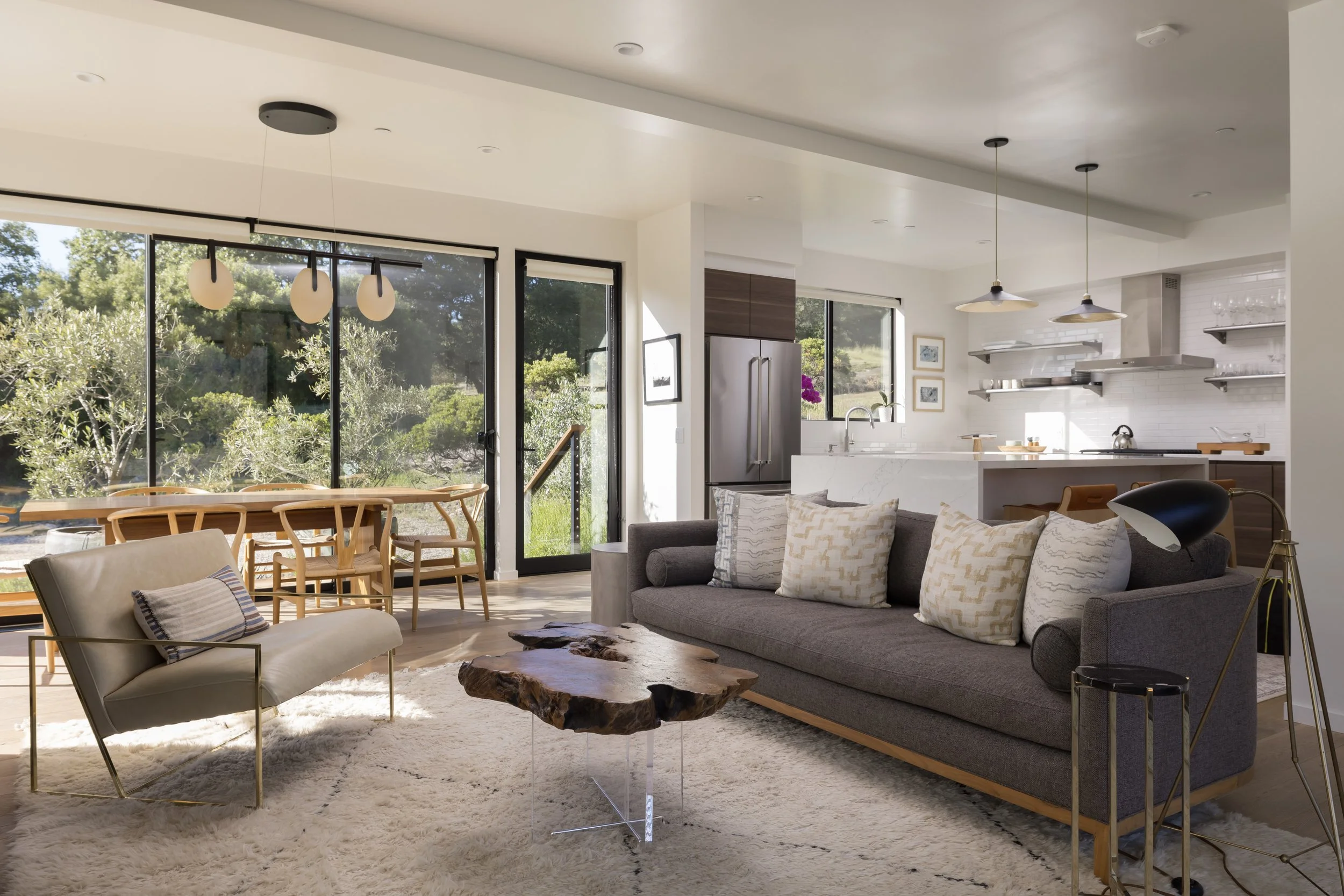 Living room with large windows, gray sofa with decorative pillows, wooden coffee table, modern light fixtures, and a view of greenery outside.