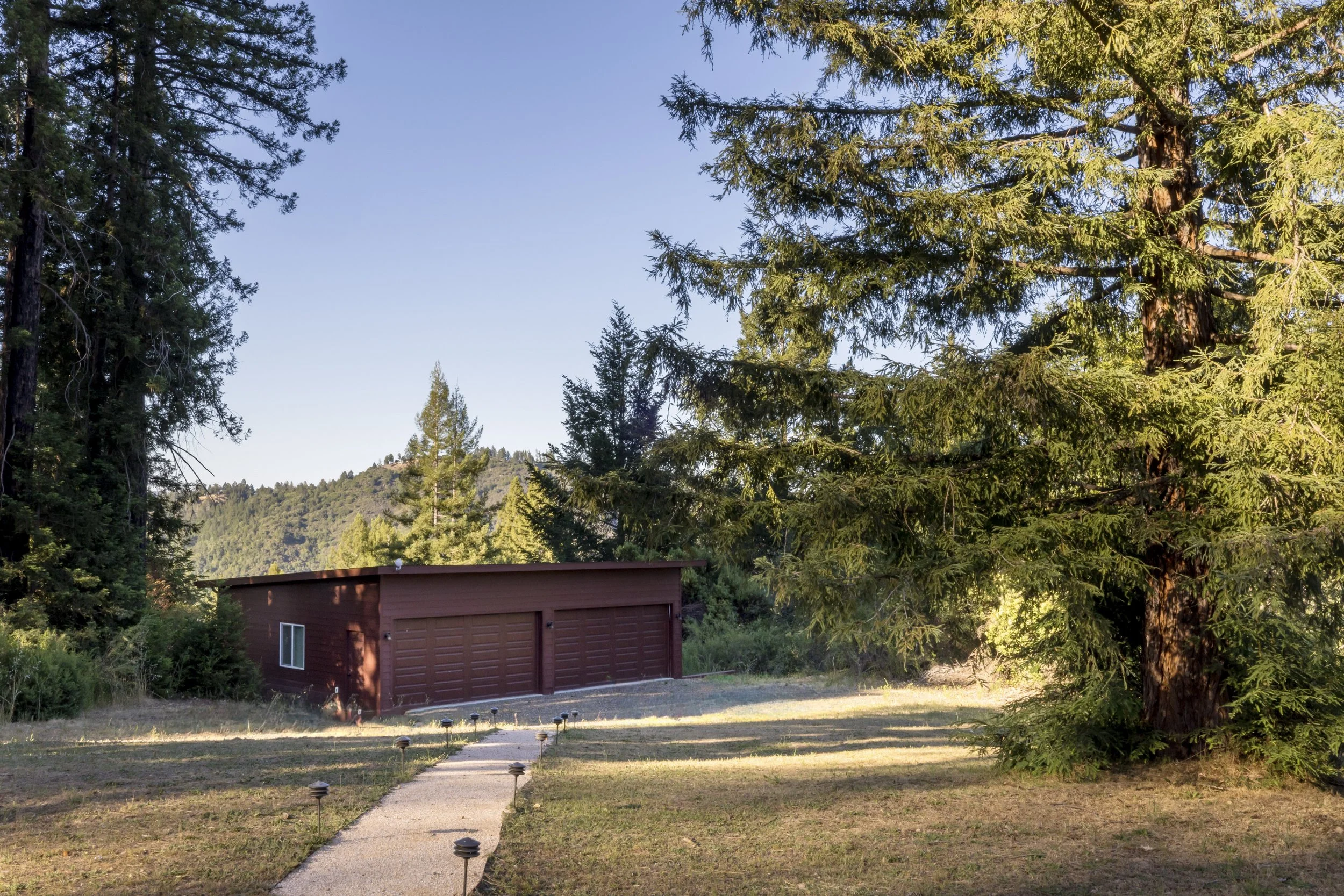 A red barn with a garage door and window situated among tall pine trees, with a mountain and blue sky in the background.