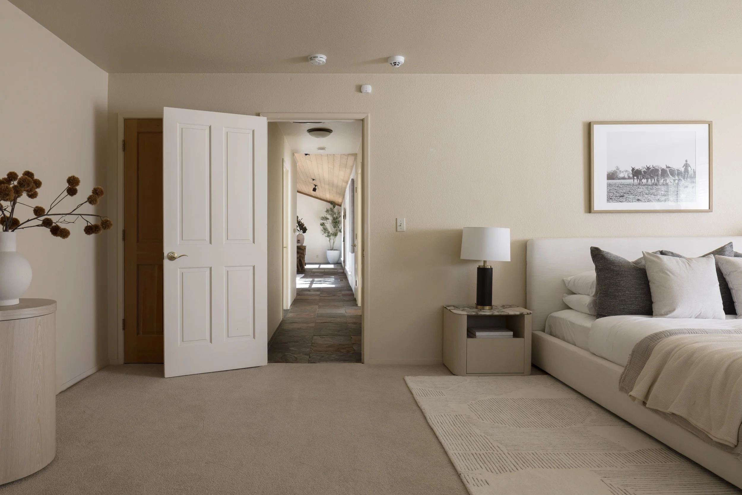 Interior of a living room with beige walls, a white sofa with pillows, a small side table with a lamp, a framed black and white photo of cows on the wall, and a doorway leading to a hallway with plants and wood ceiling.