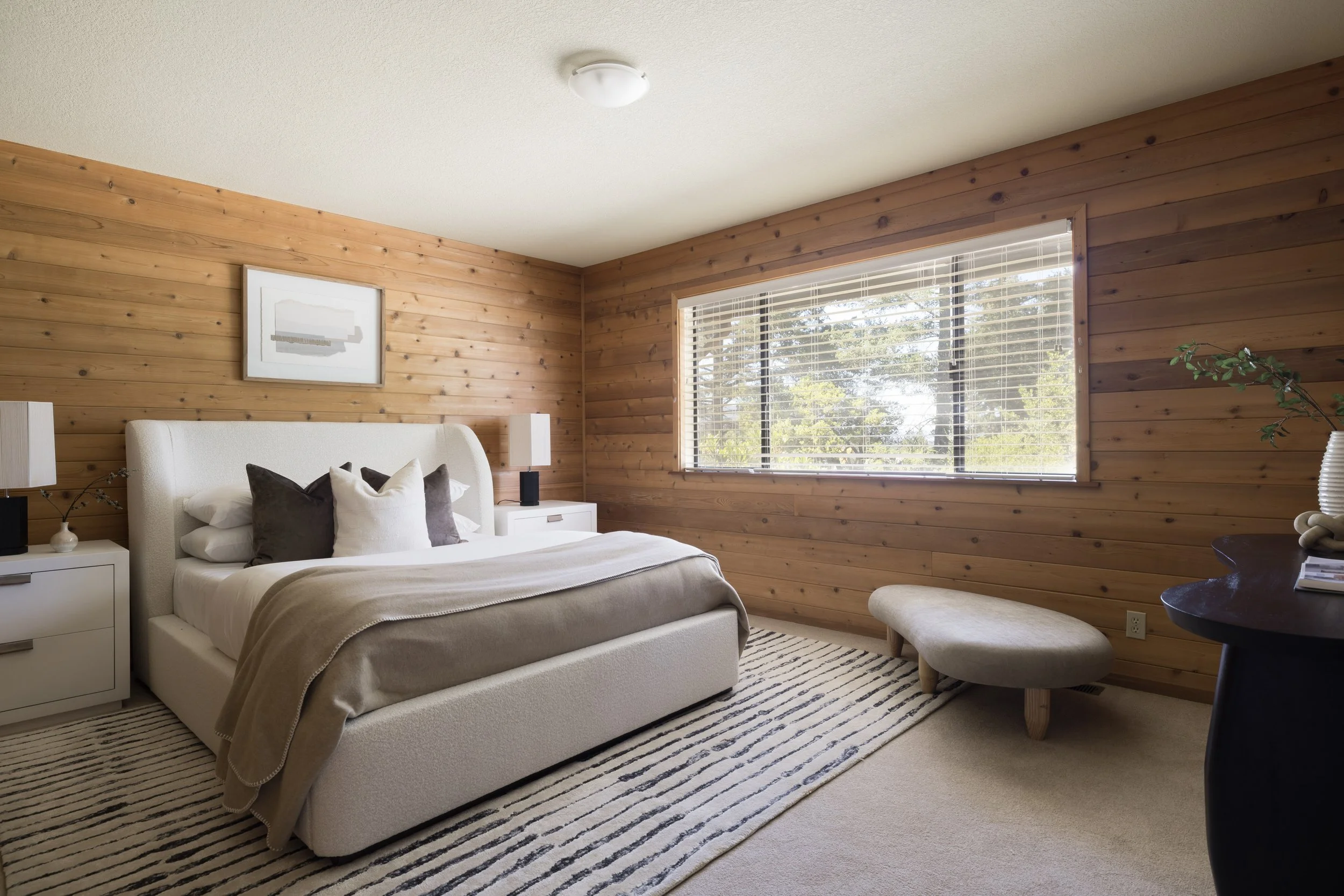 A bedroom with wood paneled walls, a white bed with pillows, a window with blinds, and a black side table with a plant and books.