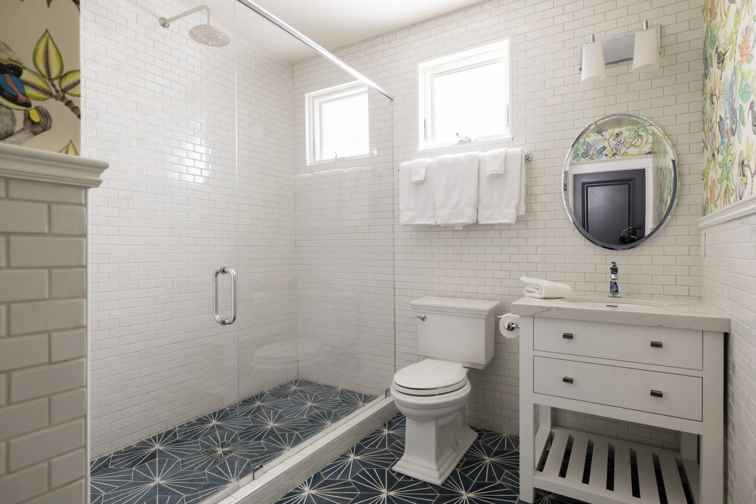 A bathroom with white brick walls, a glass-enclosed shower area with a showerhead and a window, a white toilet, white vanity with a round mirror, a towel rack with white towels, and decorative wallpaper on one wall.