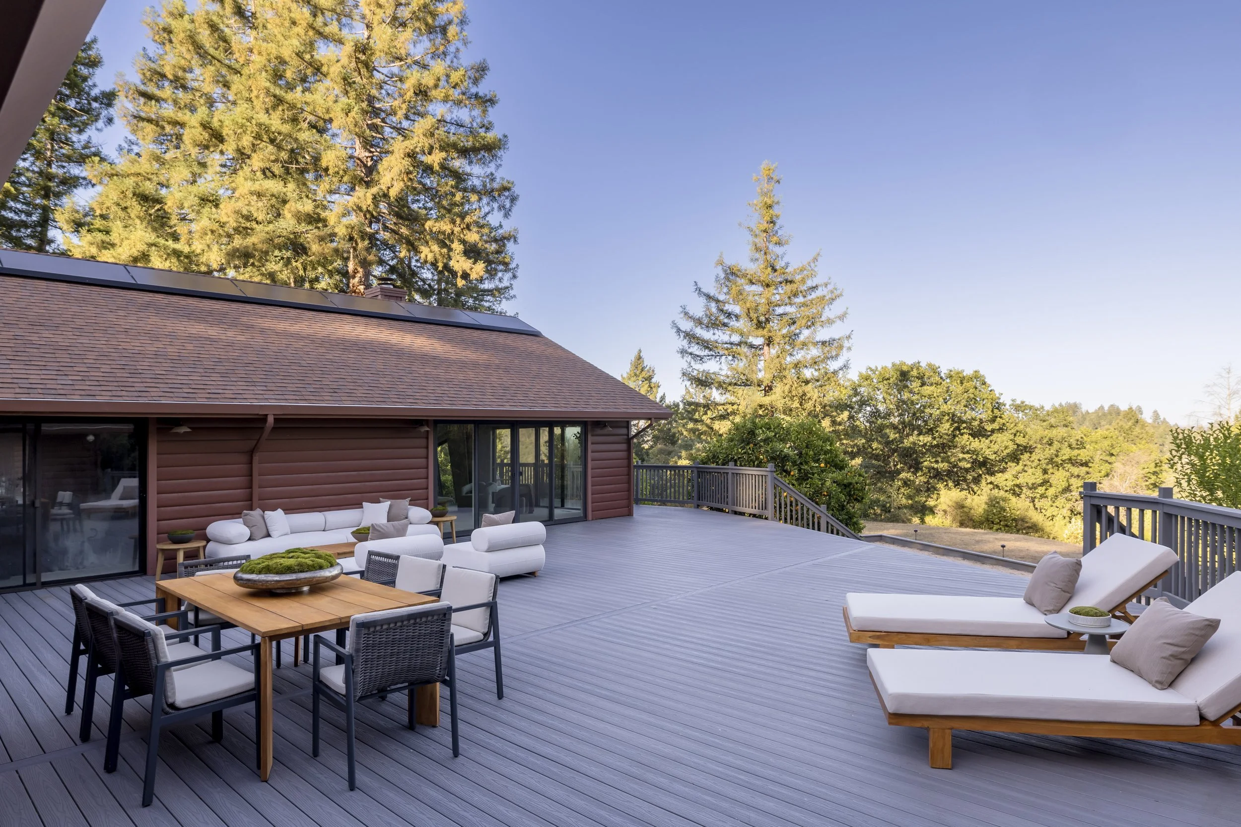 A spacious outdoor deck with modern patio furniture, including a white sofa with pillows, a wooden table with black chairs, and a seating area with cushioned lounge chairs. The deck is attached to a house with a reddish-brown exterior, large glass do