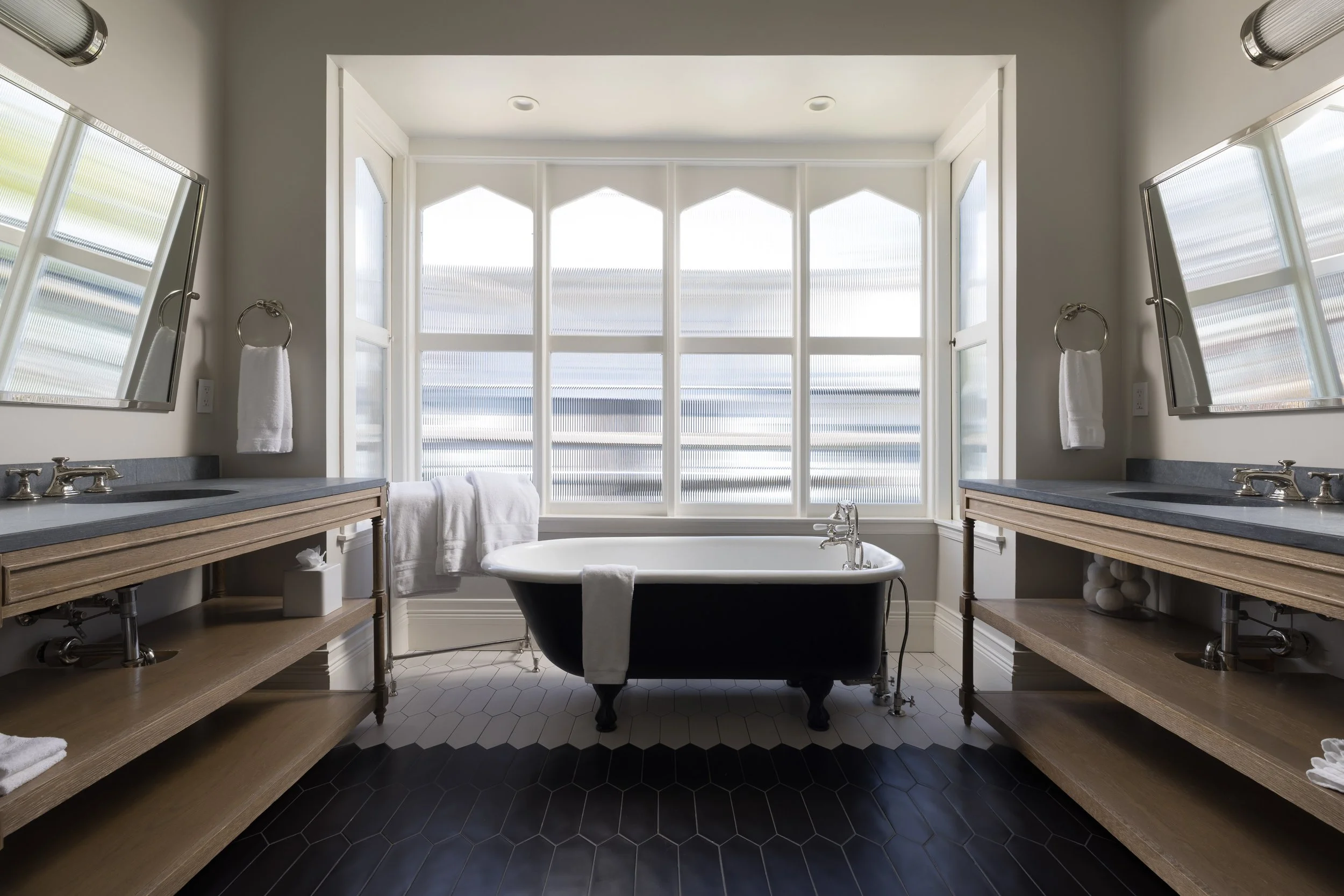 Bathroom with a black clawfoot tub centered in front of a large window, two wooden vanities with gray countertops and silver fixtures on either side, white towels hanging on silver towel rings, hexagonal black floor tiles, and two mirrors above each 