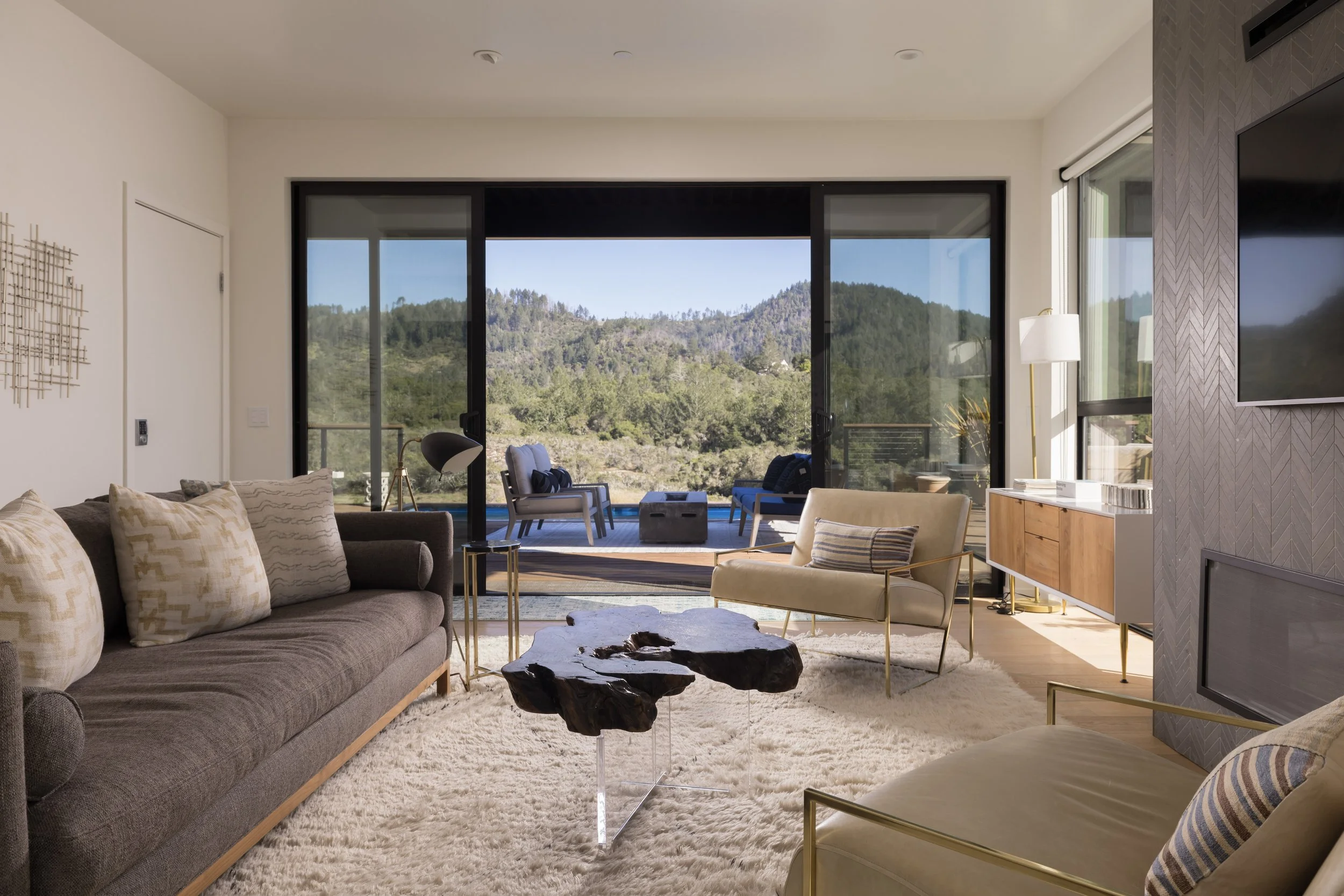 Living room with beige and brown furniture, large sliding glass doors opening to a balcony with mountain view, and a flat-screen TV on a patterned wall.