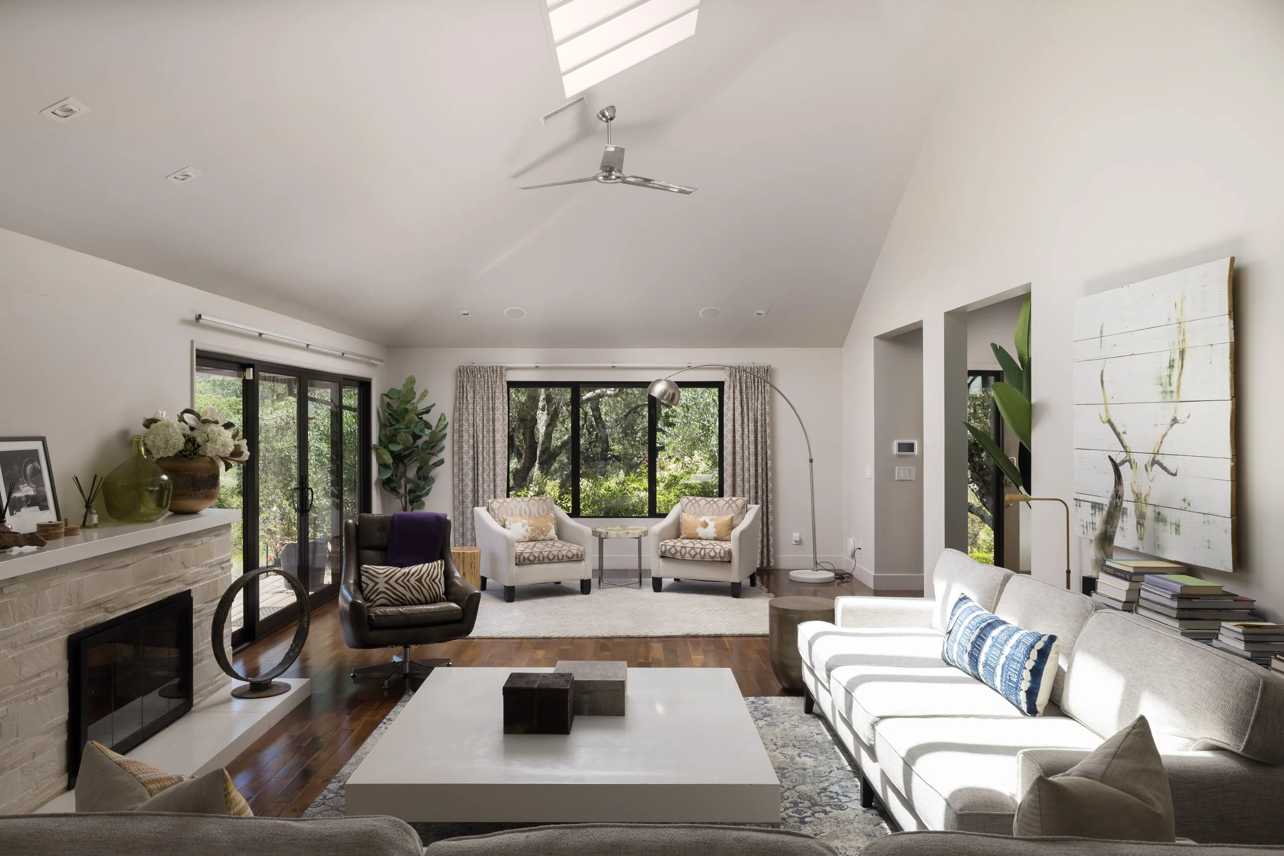 Bright living room with white walls, large windows, and ceiling fan. Furniture includes a white couch with blue pillow, black armchair, and two armchairs near windows. Decor includes a white fireplace with vases and photo frame, large abstract painti