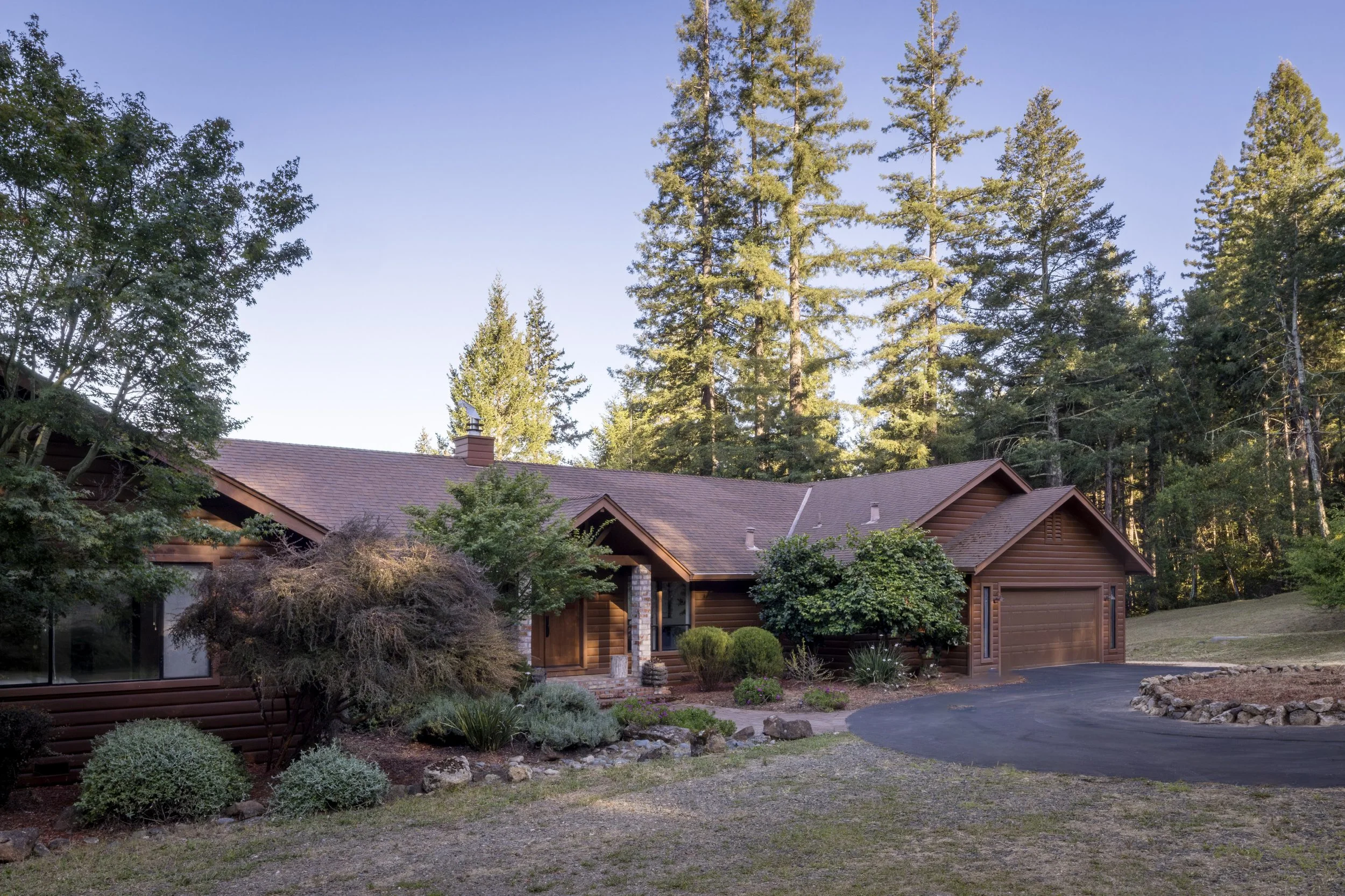 A rustic wooden house with a brown roof is set against a backdrop of tall pine trees and a clear blue sky. The front yard features landscaped bushes, small plants, and a curved driveway.