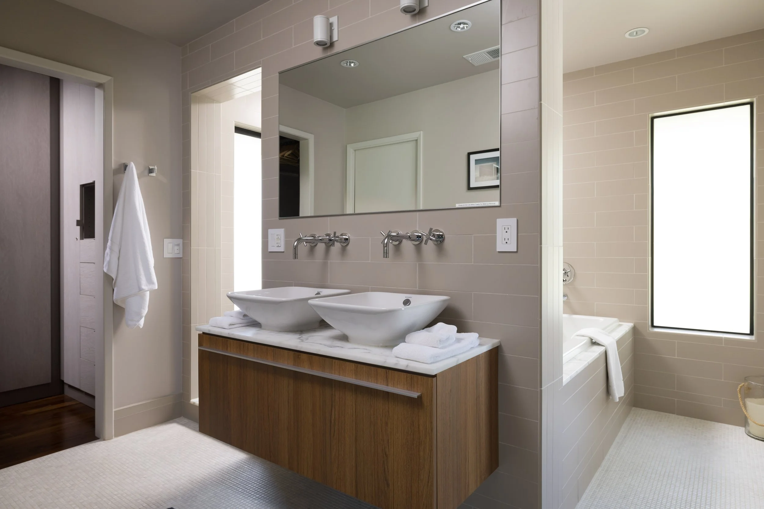 Modern bathroom with two vessel sinks on a wooden vanity, large mirror, neutral tile walls, and a window with natural light.