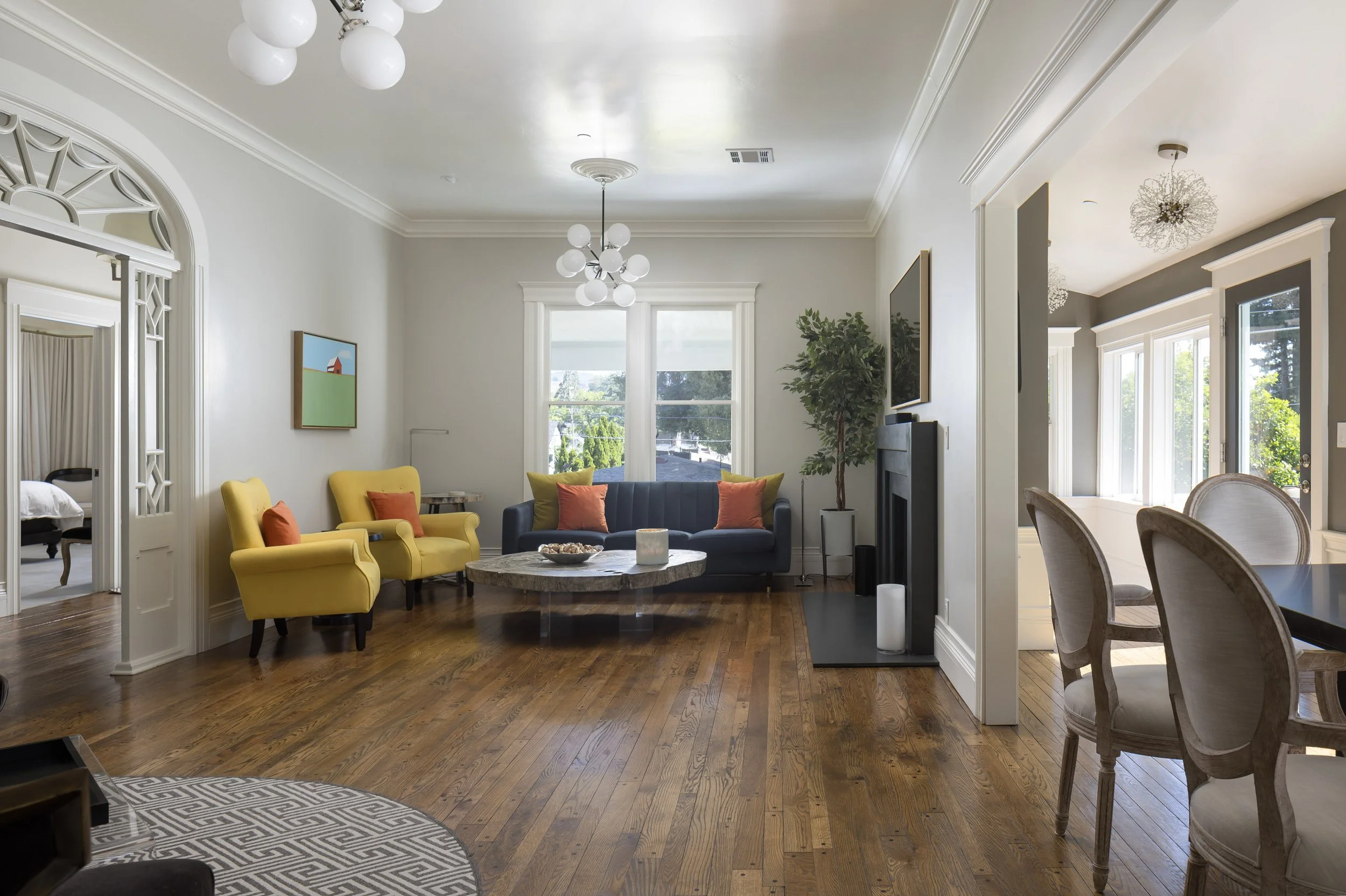Living room with yellow, blue, and gray sofas, a round coffee table, large windows, crown molding, two chandeliers, and wooden flooring.