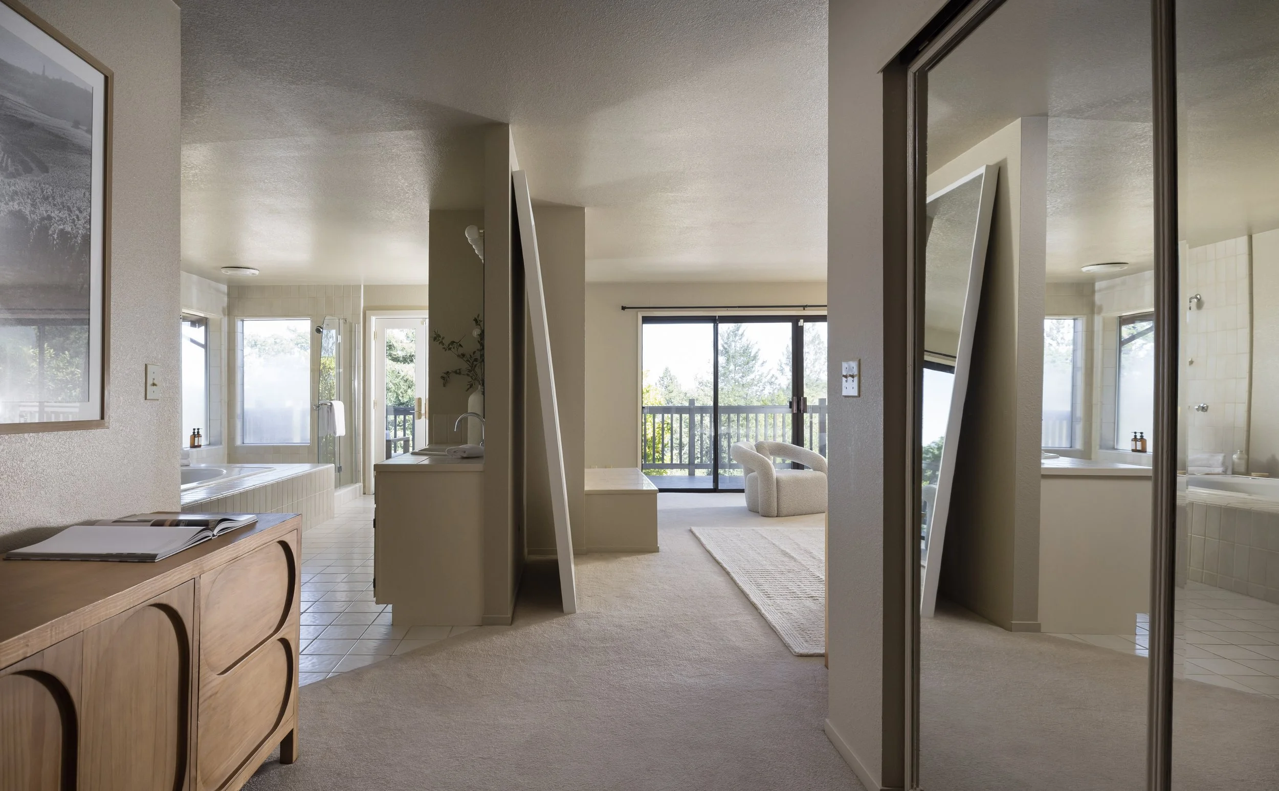 Interior view of a modern living space with a bathroom, view outside to a balcony, and a mirror reflecting part of the room. There is a wooden sideboard, a picture on the wall, and open light-colored curtains.