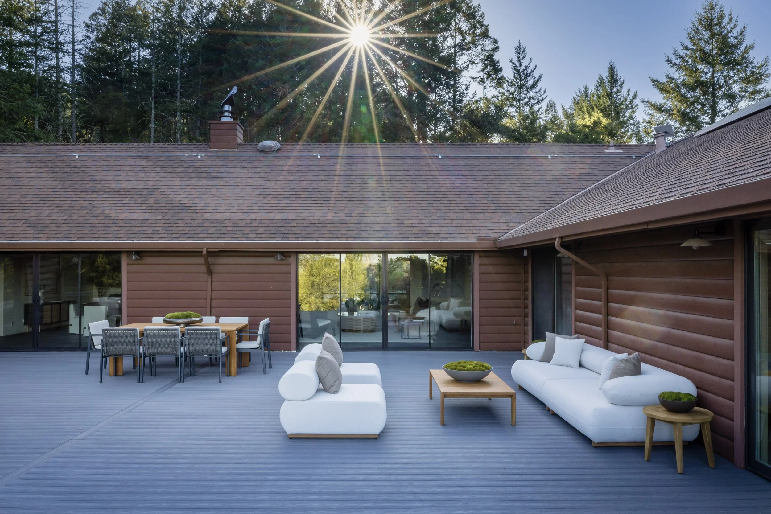 Outdoor deck with white cushioned seating, a wooden dining table with chairs, and a view of trees and a house roof under a clear sky with the sun shining.