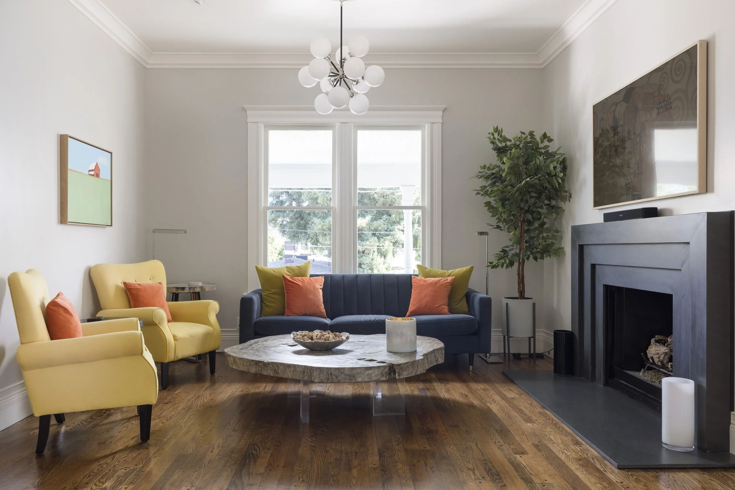 Modern living room with yellow and dark blue armchairs and colorful cushions, a round marble coffee table with a bowl of shells, a large window with natural light, framed artwork on the walls, a tall potted plant, and a black fireplace with candles a