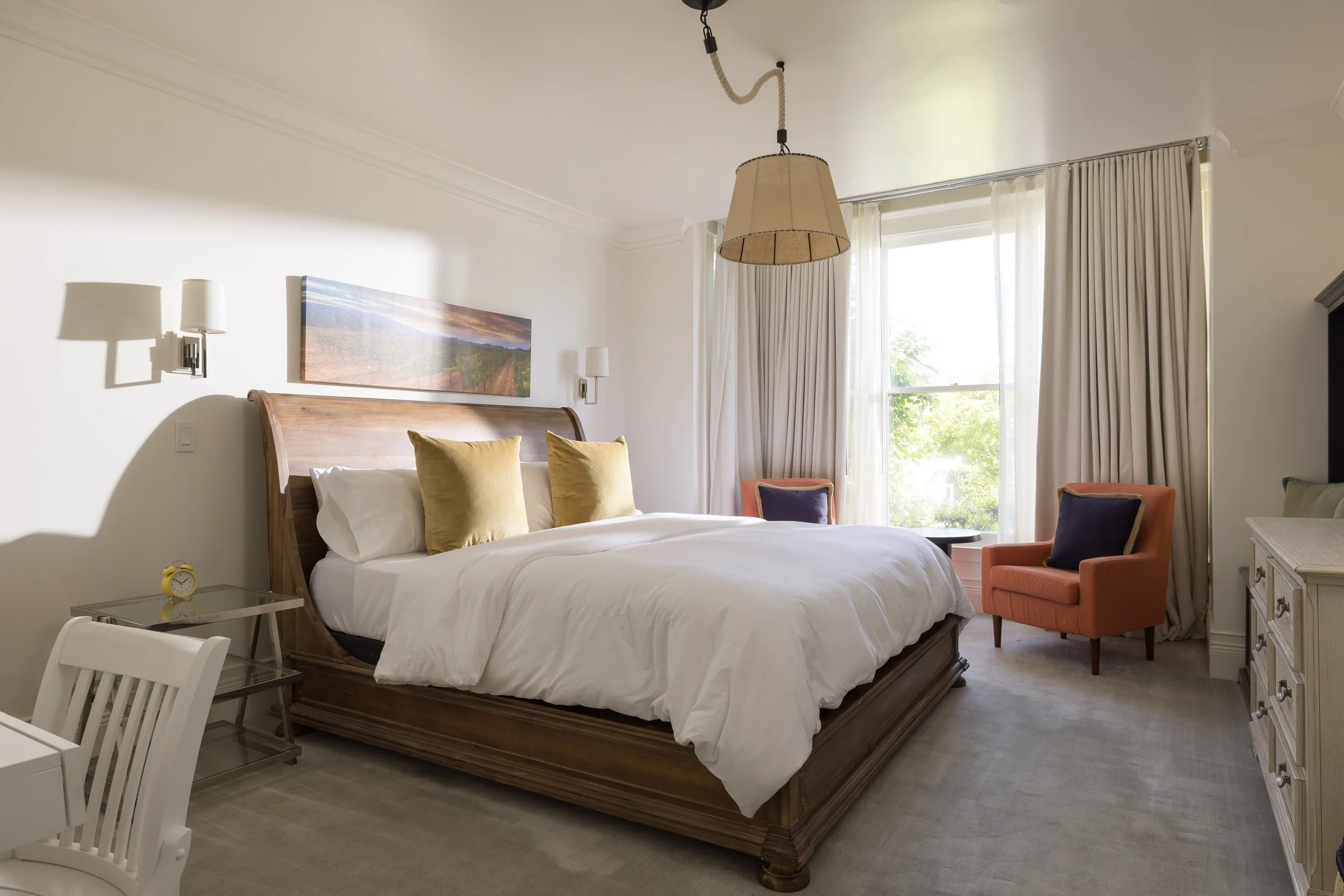 A bedroom with a wooden bed, white bedding, and yellow pillows, beside a small glass nightstand with a yellow alarm clock. There is a chair with an orange cushion and a purple pillow near a window with cream curtains, allowing sunlight into the room.