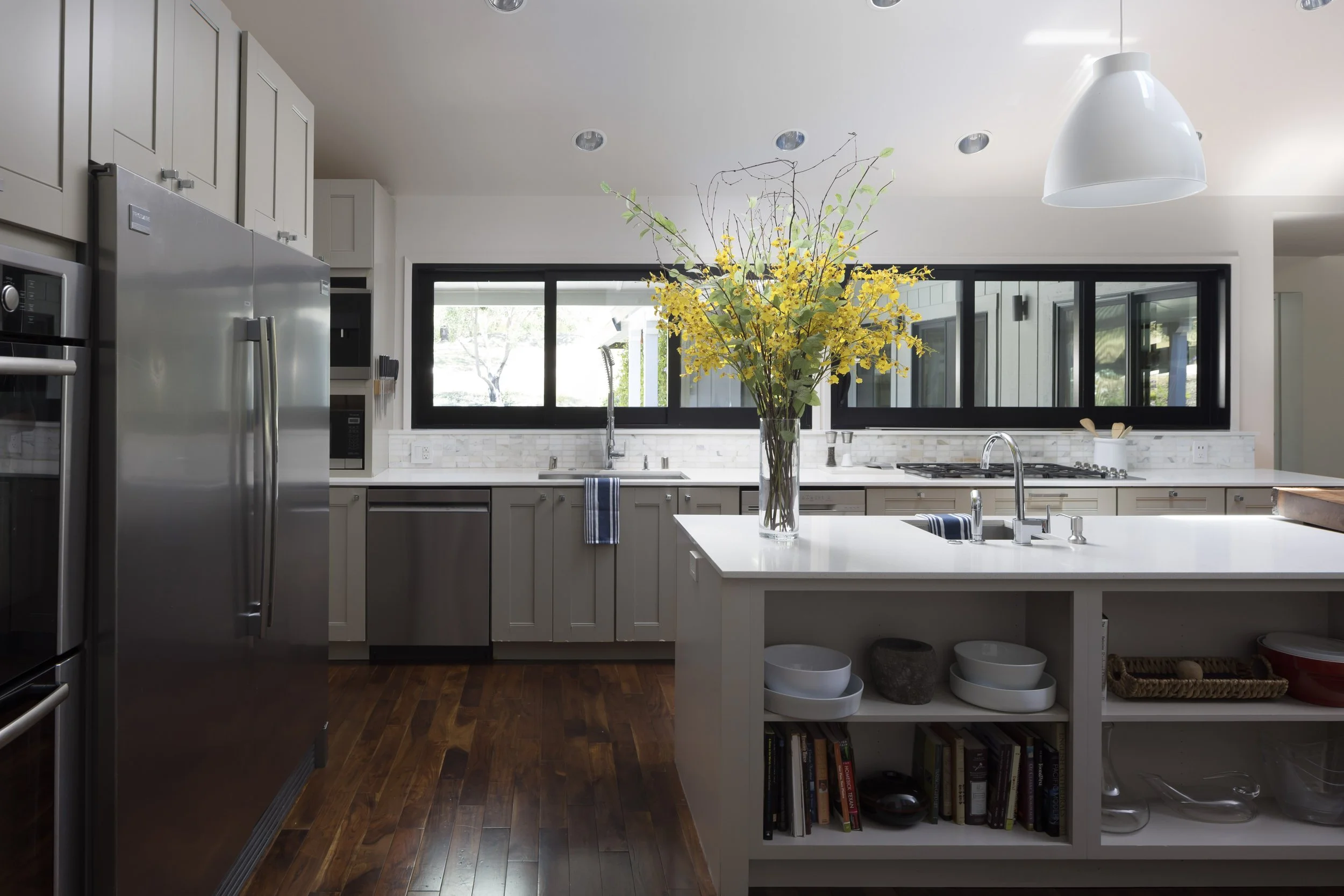 Kitchen with white cabinets, stainless steel appliances, a large window, and a white island with open storage. A vase with yellow flowers is on the island.