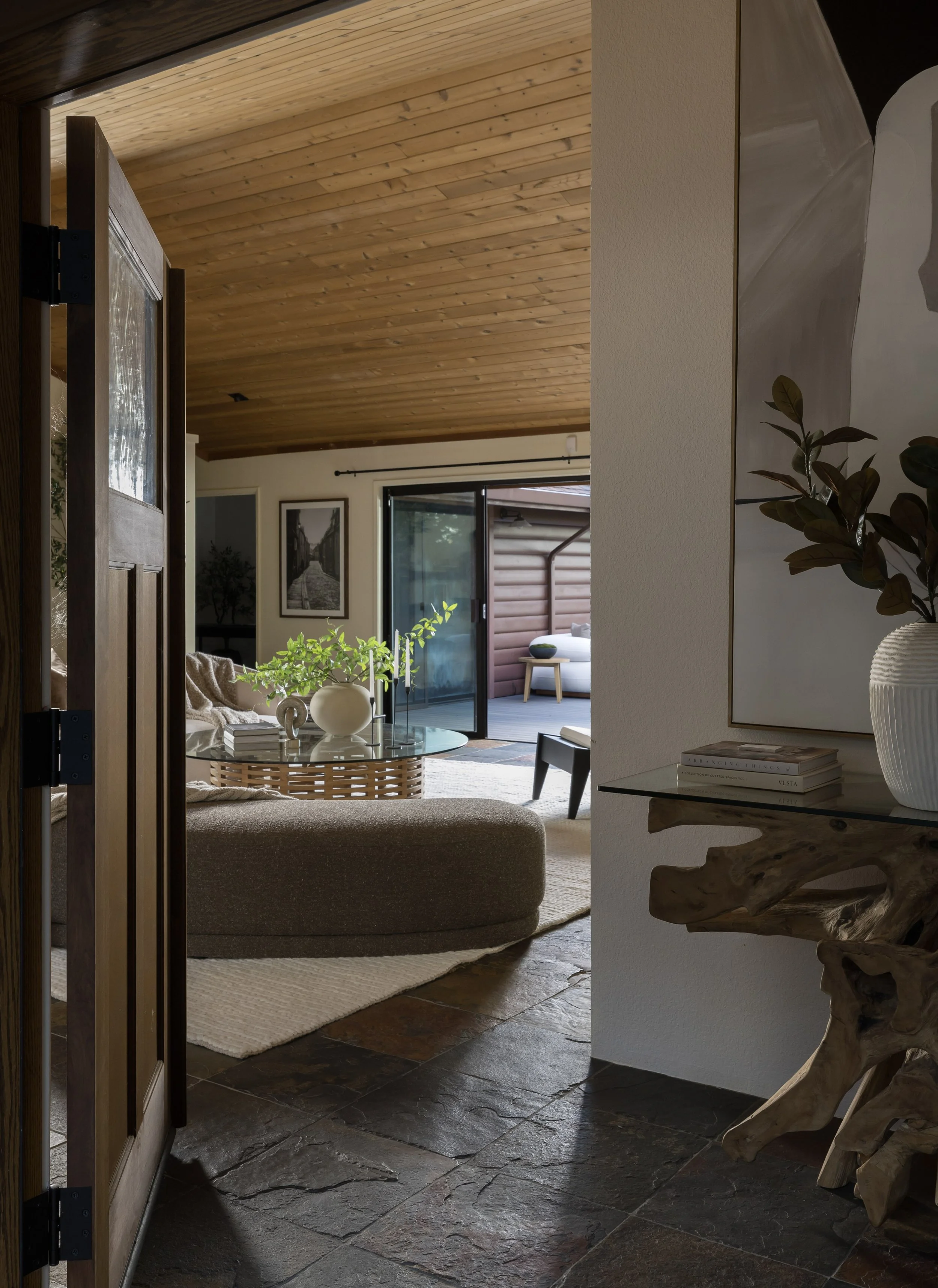 Interior living room with wooden ceiling, sliding glass door leading outside, framed black-and-white photograph, houseplants, and furniture on a rug.