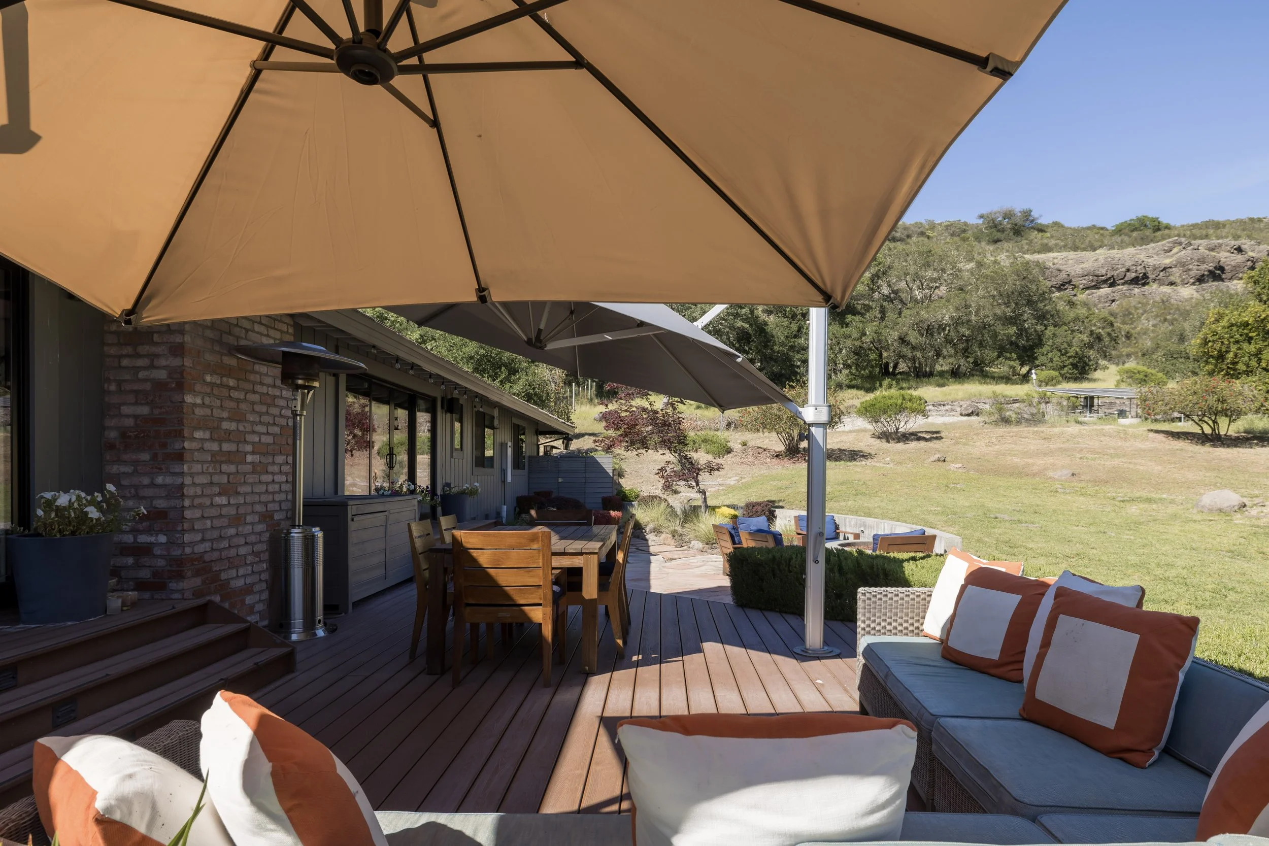 A backyard patio with large beige umbrellas shading a wooden dining table, outdoor chairs, and a cushioned lounge chair with orange and white pillows. There is a house with brick and siding and outdoor heaters, with a landscape of trees, grass, rocks