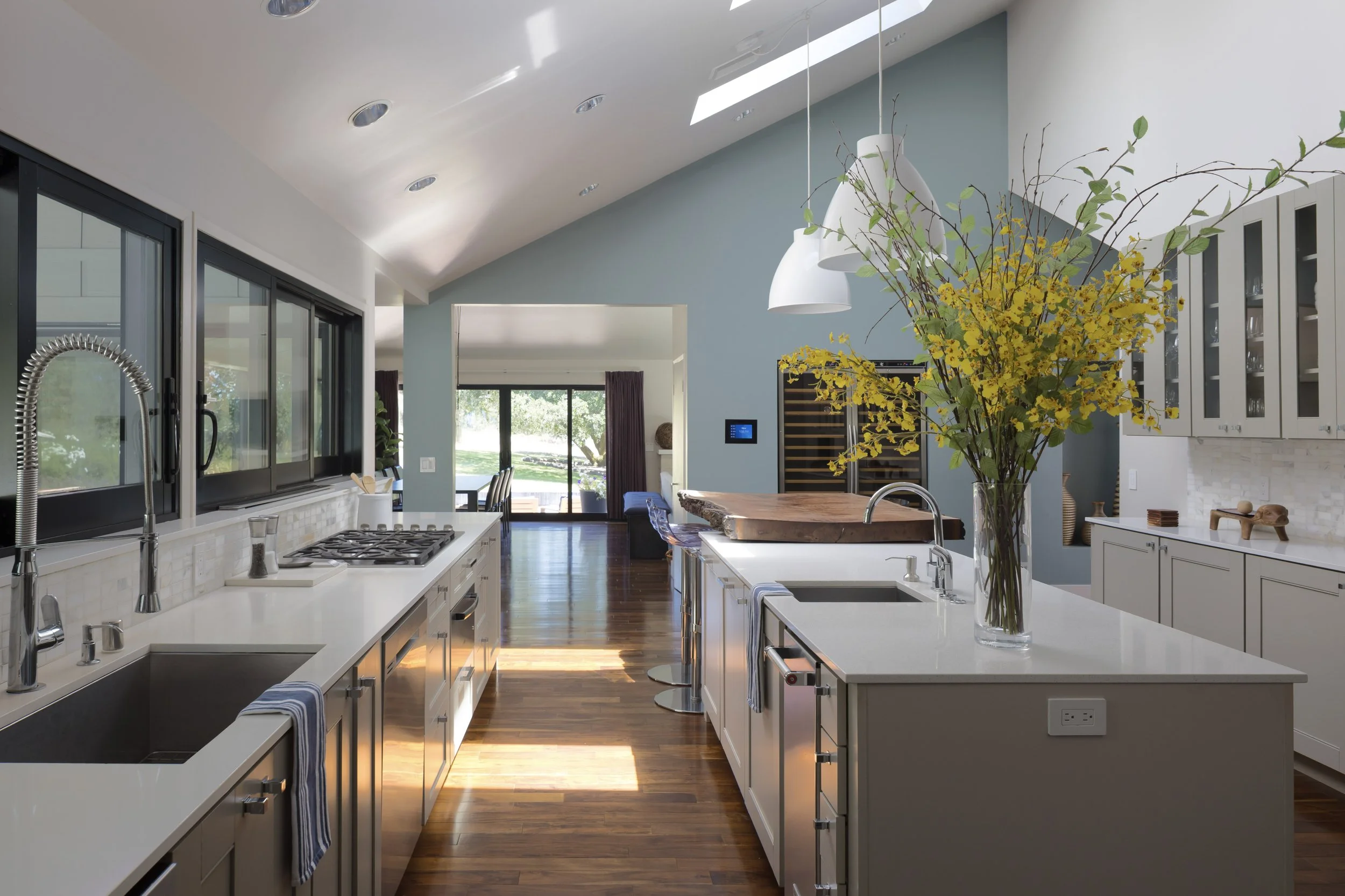 Bright kitchen with white cabinets, a large island with a vase of yellow flowers, and windows showing green outdoor scenery.