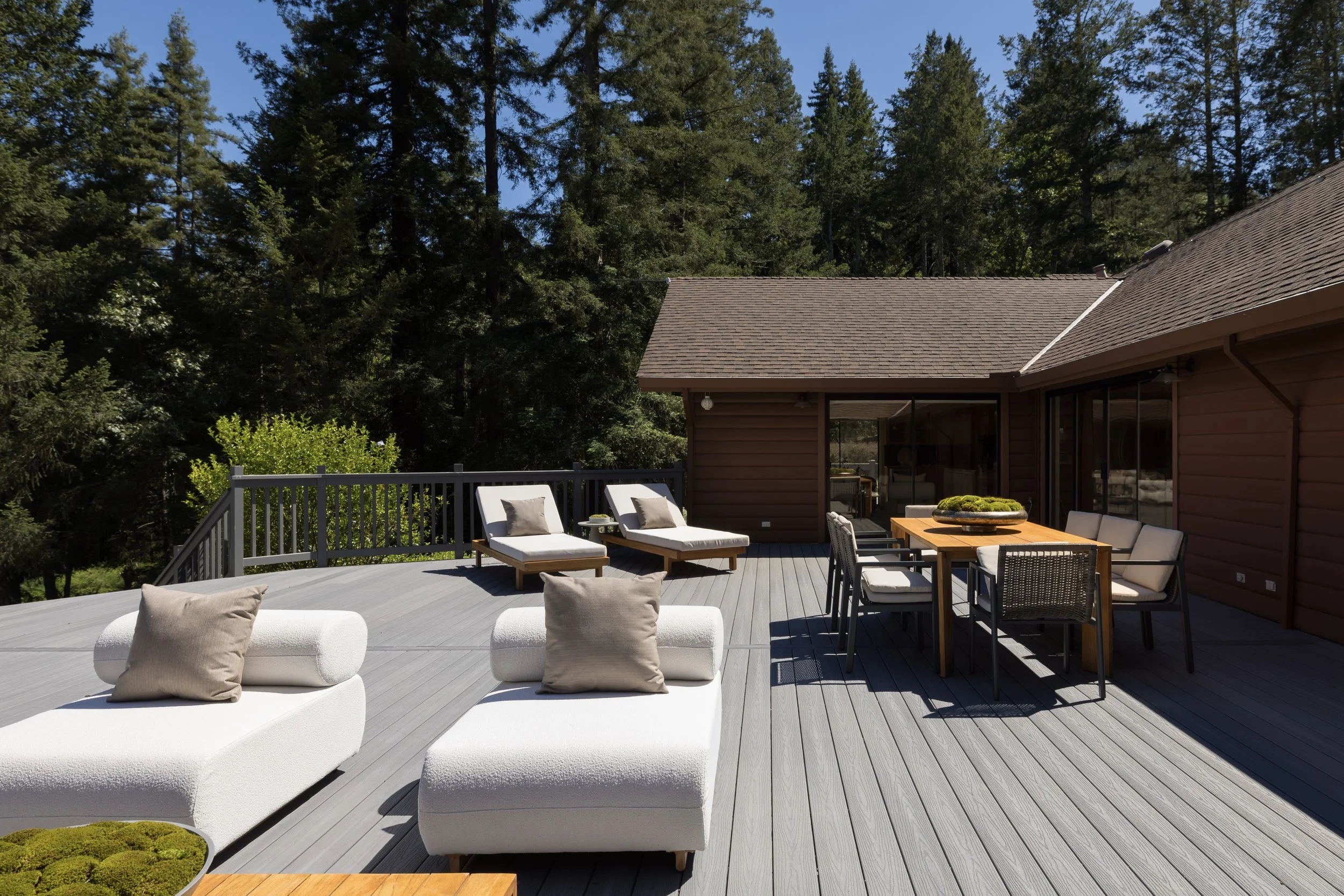 A spacious outdoor deck with white lounge chairs and a dining table with chairs, bordered by a grey railing, with lush green trees and a house in the background under a clear blue sky.