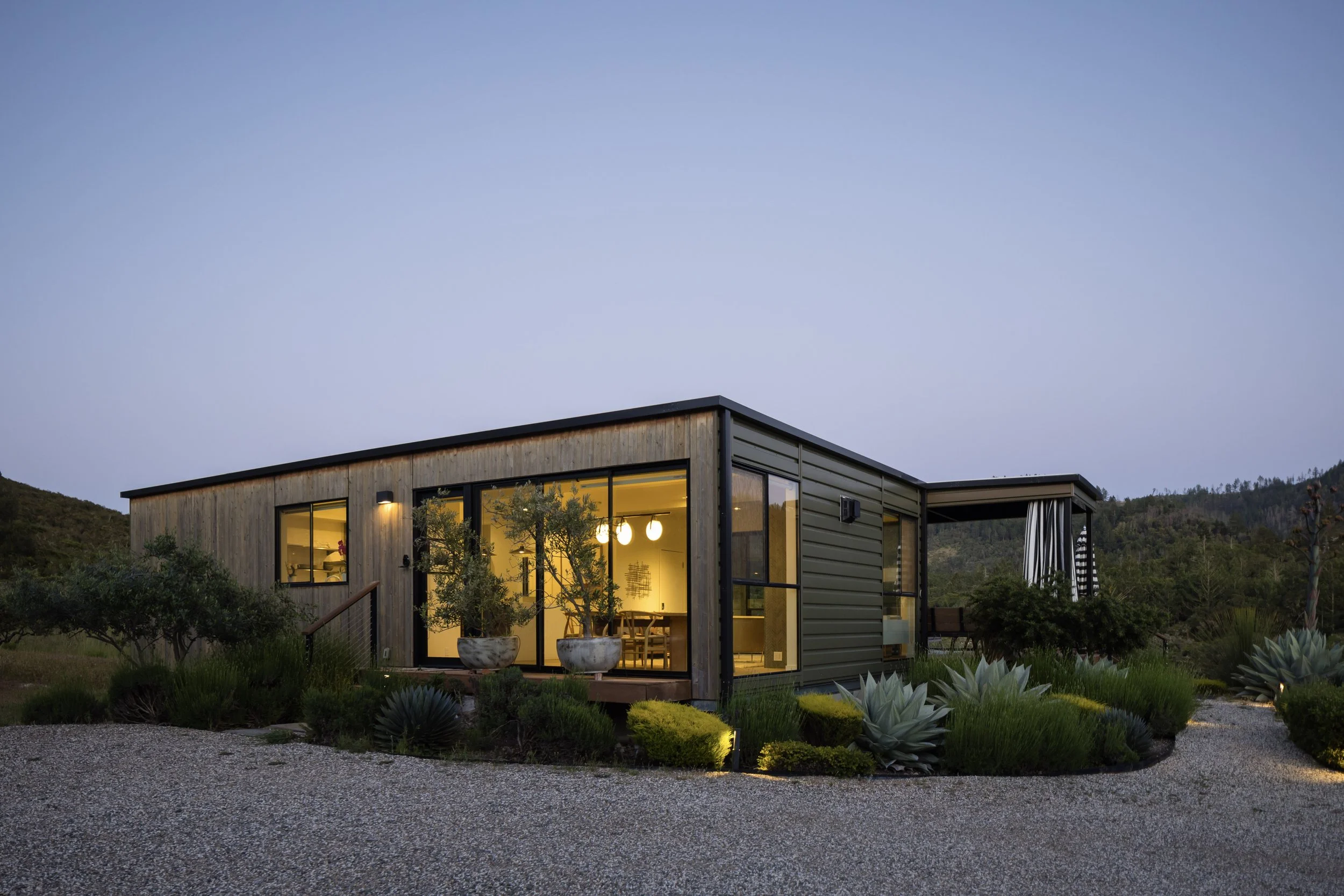Modern house with large glass windows and warm interior lighting, situated in a landscaped garden with various plants and bushes, under early evening sky.