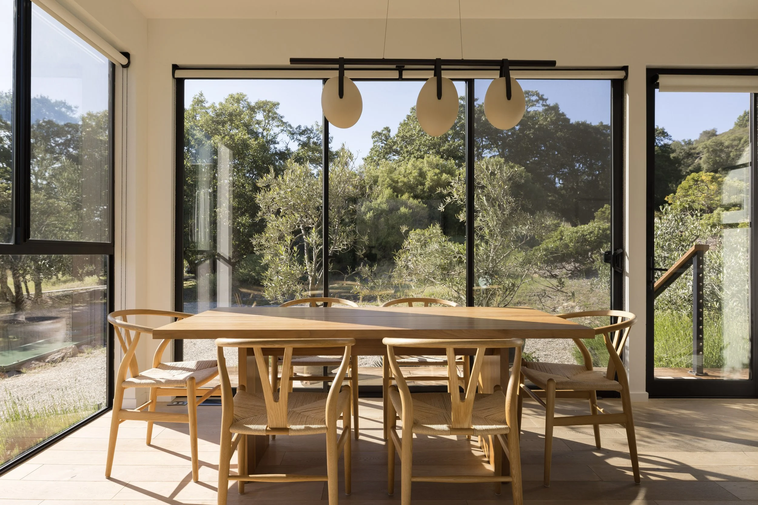 A dining room with a wooden table and chairs, large glass windows showing outdoor greenery, and a modern hanging light fixture.