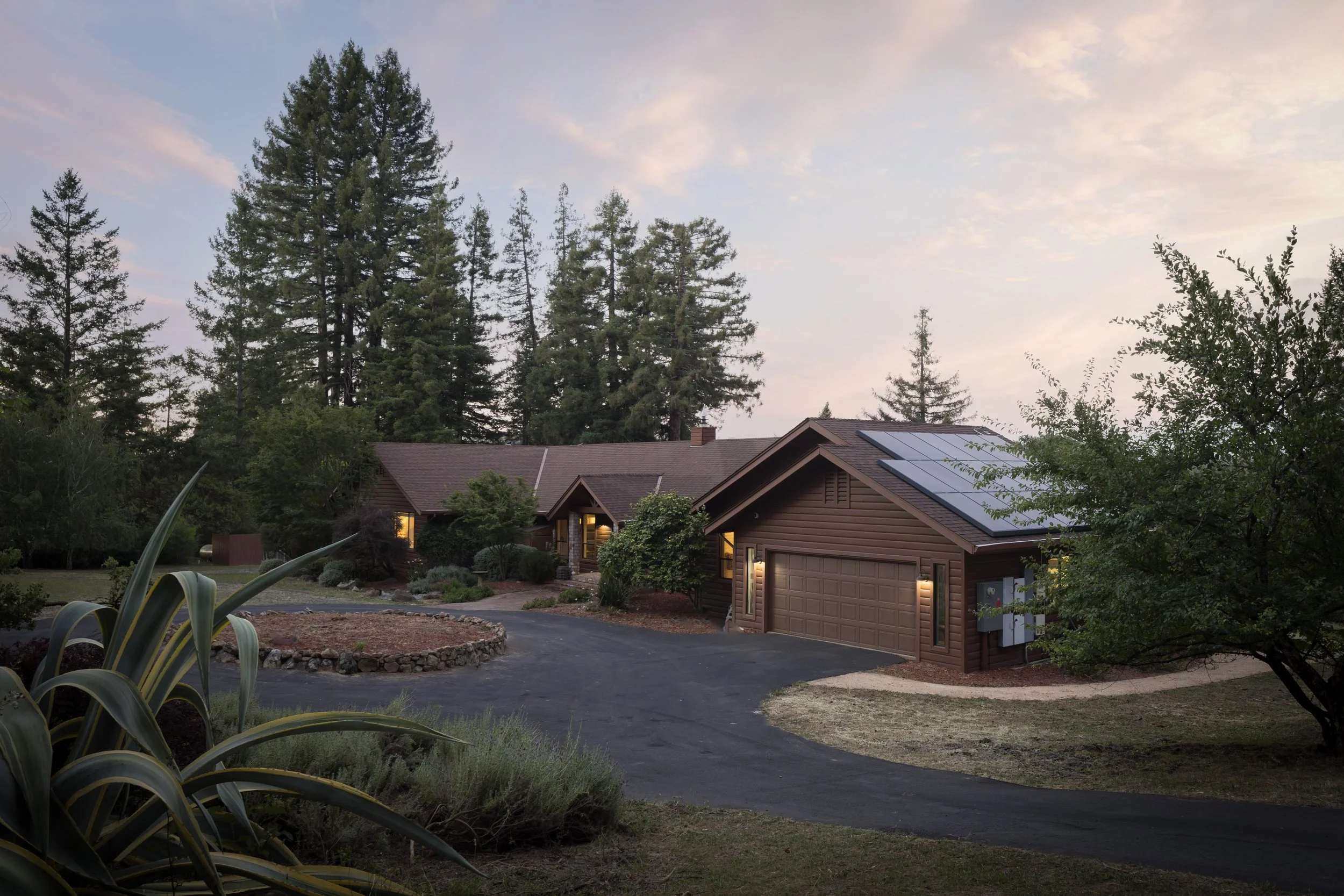 A house with a brown roof and solar panels, surrounded by trees and landscaping, at dusk with the sky partly cloudy.