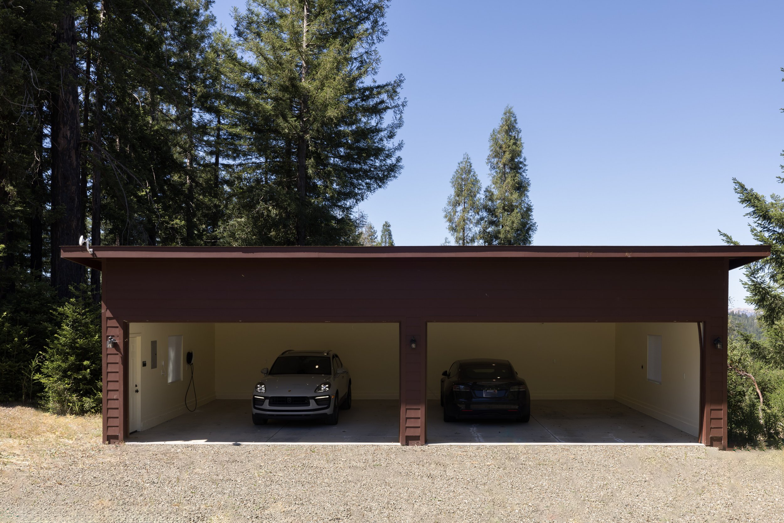 A two-car garage with a red exterior and open doors. Inside the garage, there are two cars parked side by side, one white and one black. The background features tall green pine trees under a clear blue sky.