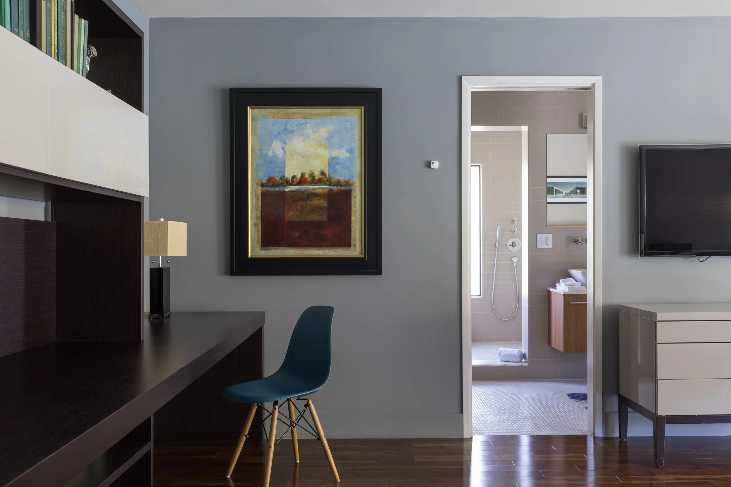Interior view of a room with a dark wooden desk, blue chair, wall art, and a visible bathroom through an open doorway.
