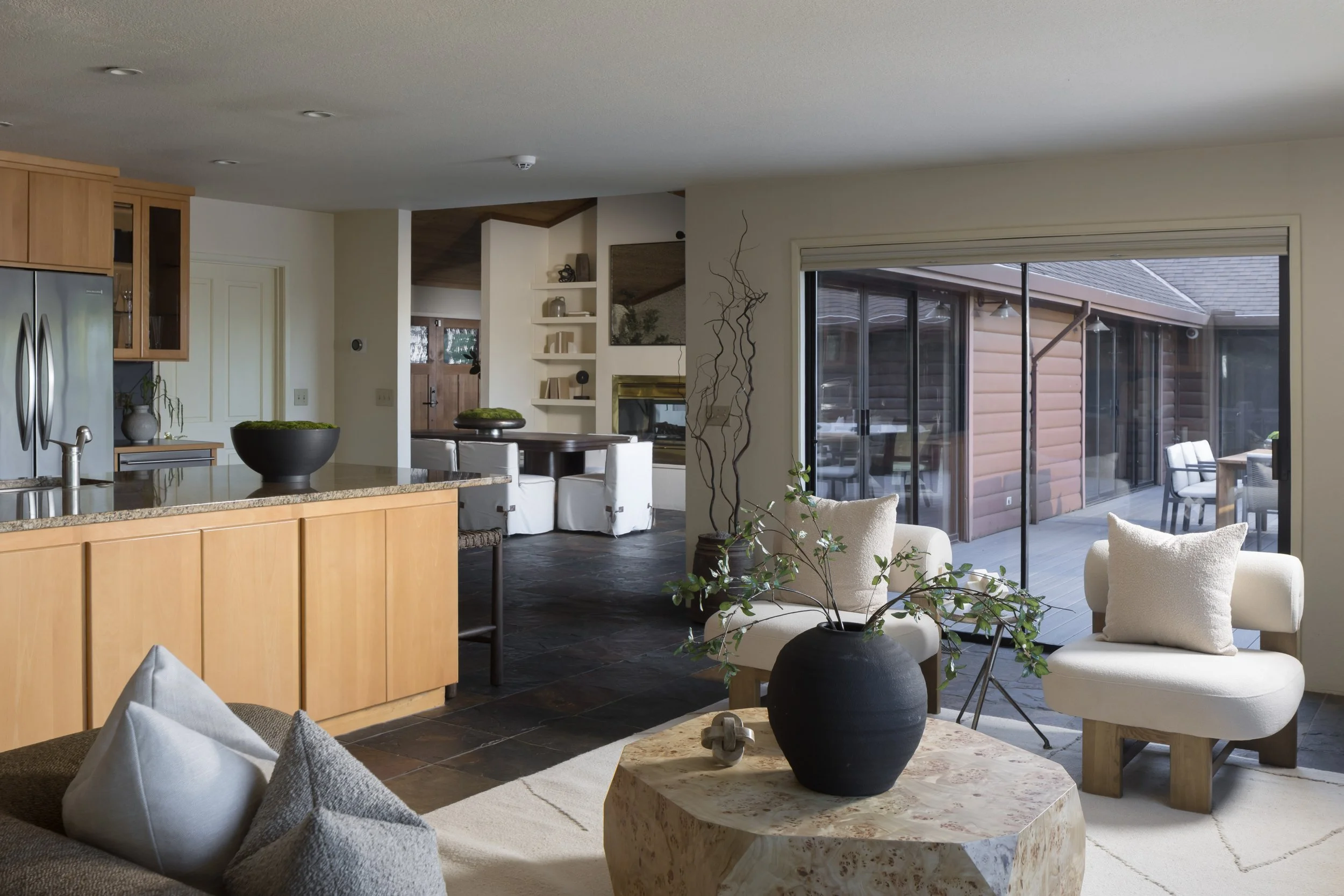 A modern living room with a view of a deck outside through sliding glass doors. The room has white seating with cushions, a black vase with a green plant on a marble table, and wooden accents. There are built-in shelves with decorative items and a fi