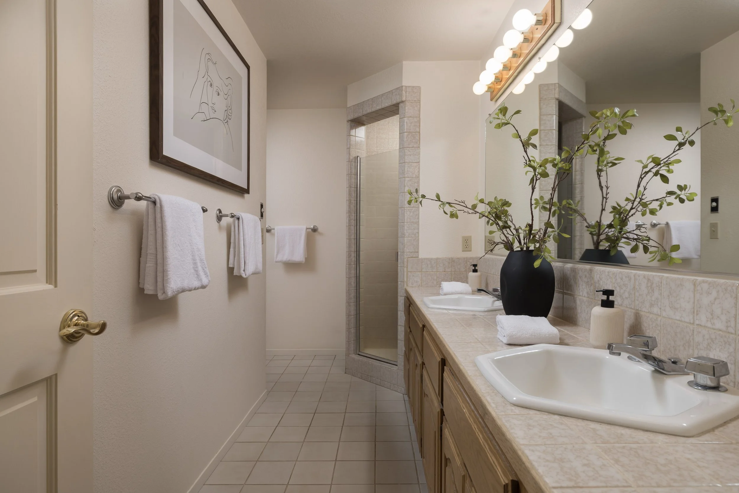 A bathroom with a beige countertop, a white sink, a mirror, potted plant, light bulbs above mirror, tiled shower, and towels on the wall.