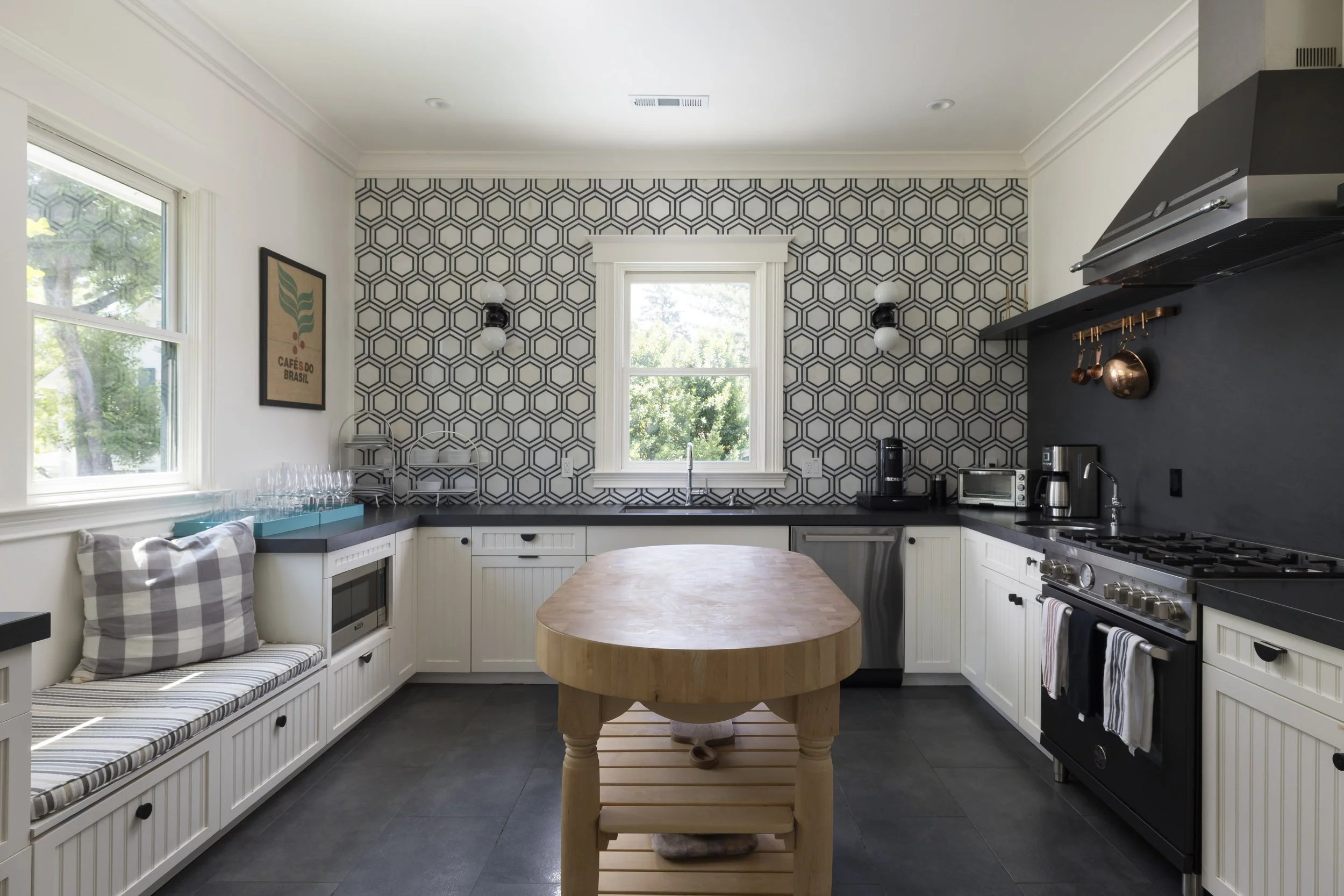 A modern kitchen with white cabinetry, geometric wallpaper, and black countertops. Features large windows with views of trees, a built-in bench with plaid cushions, and a round wooden table in the center. Appliances include a microwave, coffee maker,