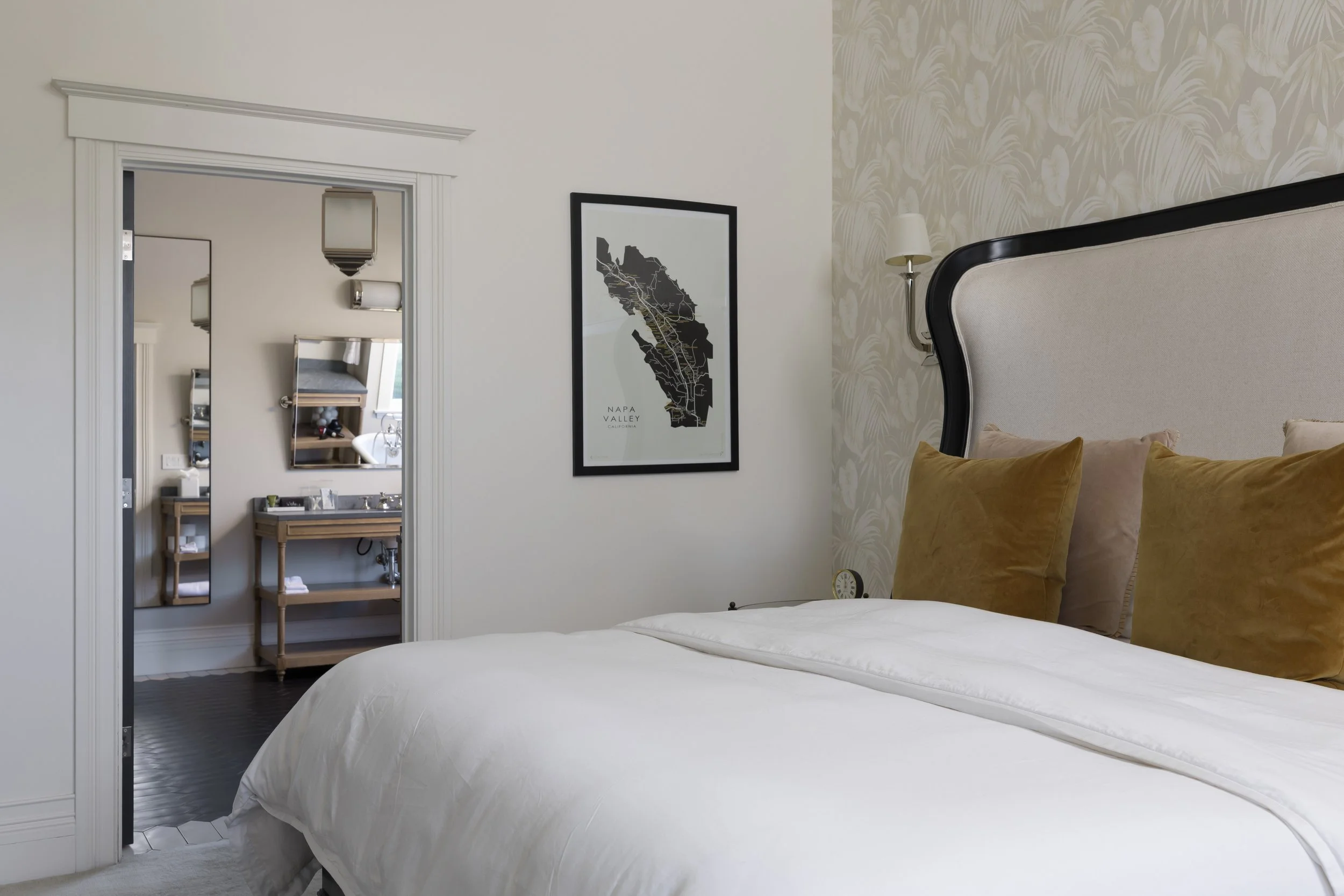 A bedroom with a bed featuring beige pillows, a wall-mounted lamp, a framed map of Napa Valley, and an adjoining bathroom area with mirrors and wooden furniture.