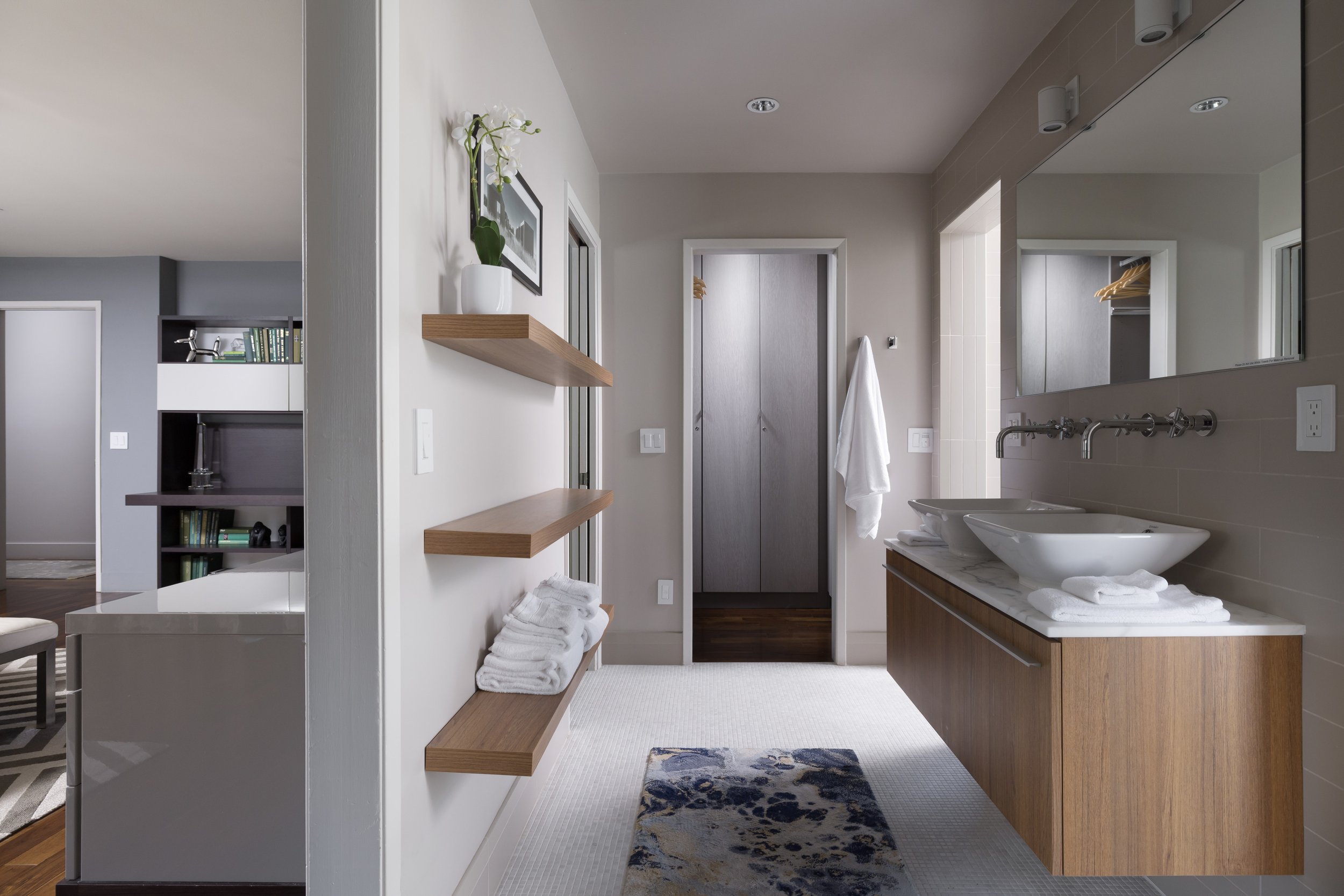 Modern bathroom with double sinks, large mirror, gray tiled walls, wooden vanity, white towels, small rug, and a closet with hanging space visible through a doorway.