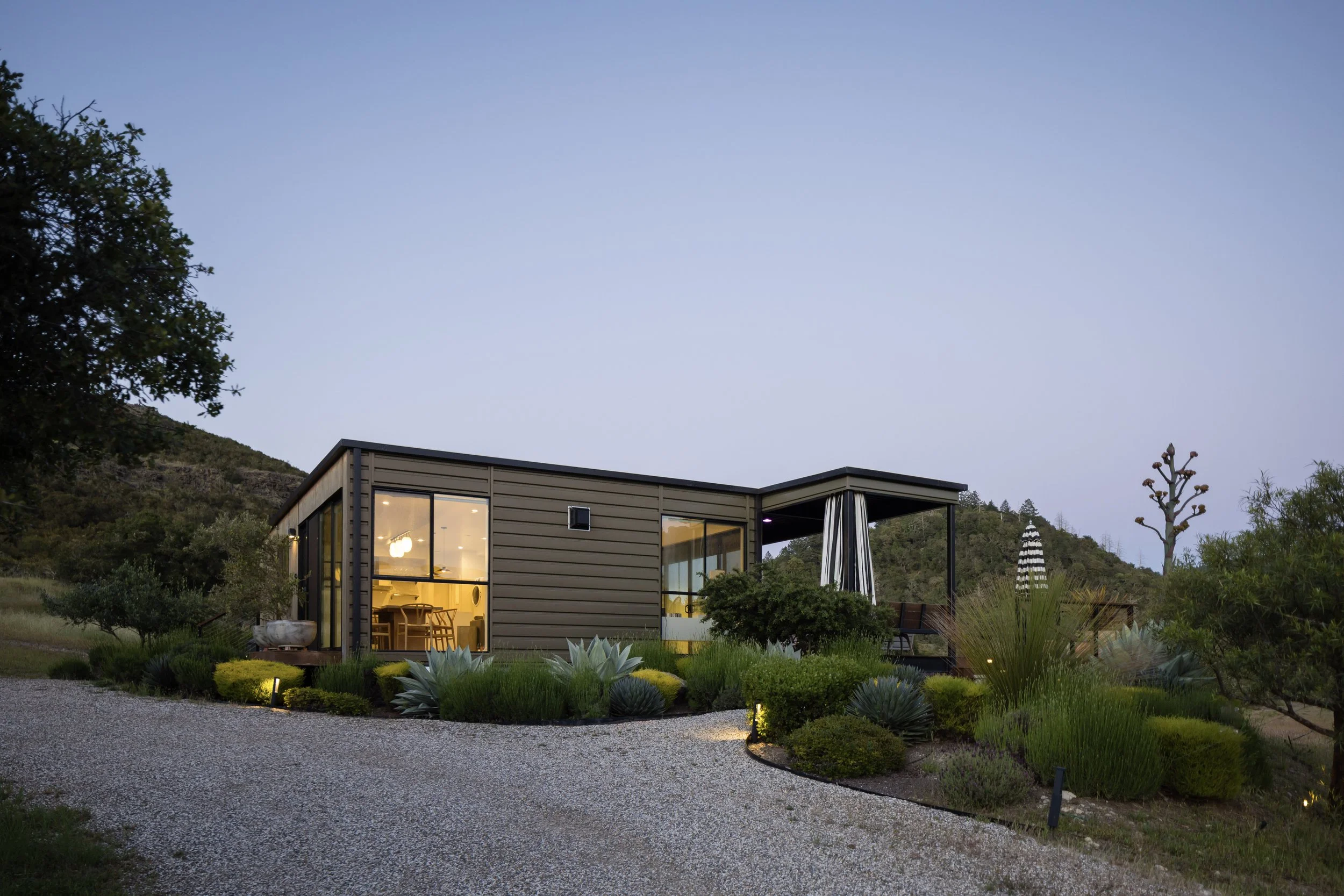 Modern house with large glass windows illuminated from inside, surrounded by desert plants and a gravel pathway, with hilly terrain and trees in the background during dusk.