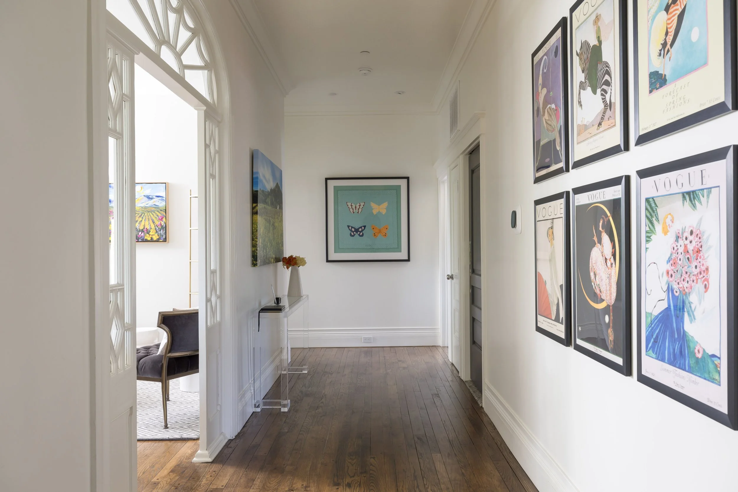 Hallway in an art gallery with framed artwork on the walls, a small table with a vase of flowers at the end, and a decorative chair near an open doorway leading to a room with more art.