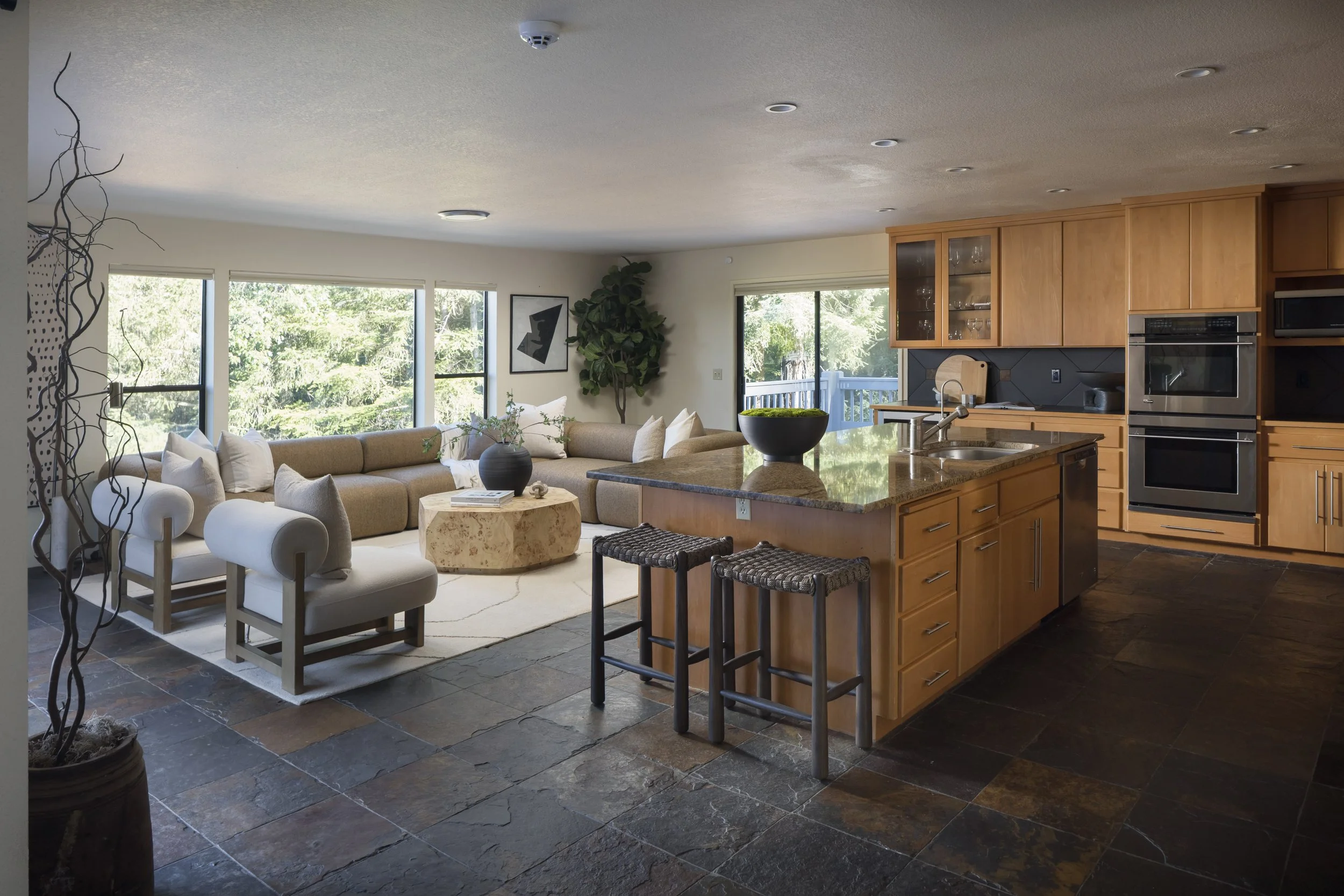 Open-concept living room and kitchen with large windows, beige sectional sofa, modern artwork, indoor plant, kitchen island with three barstools, wooden cabinets, stainless steel appliances, and tiled flooring.