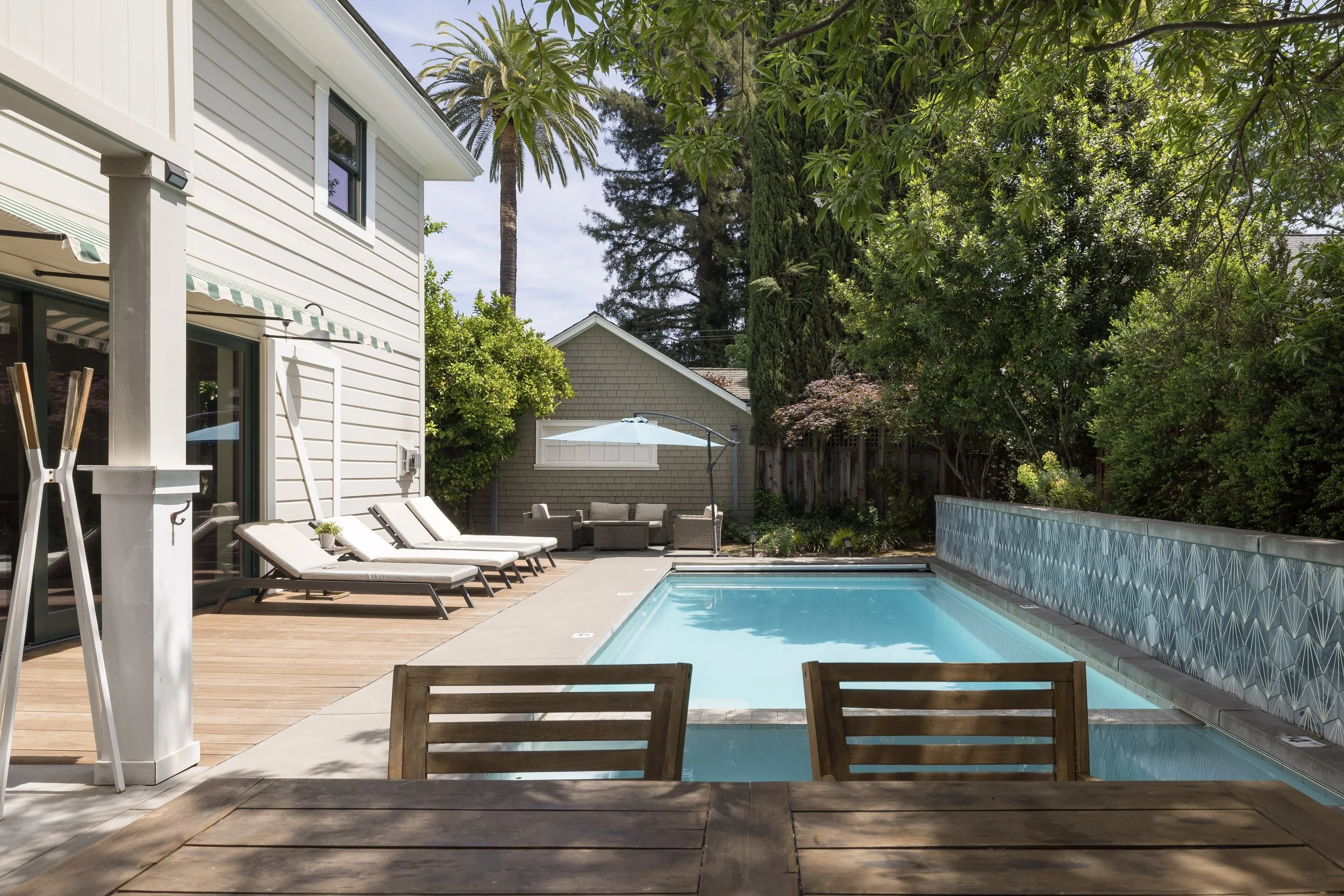 Residential backyard with a swimming pool, poolside lounge chairs, patio furniture, large trees, and a house with white siding and a green-and-white striped awning.